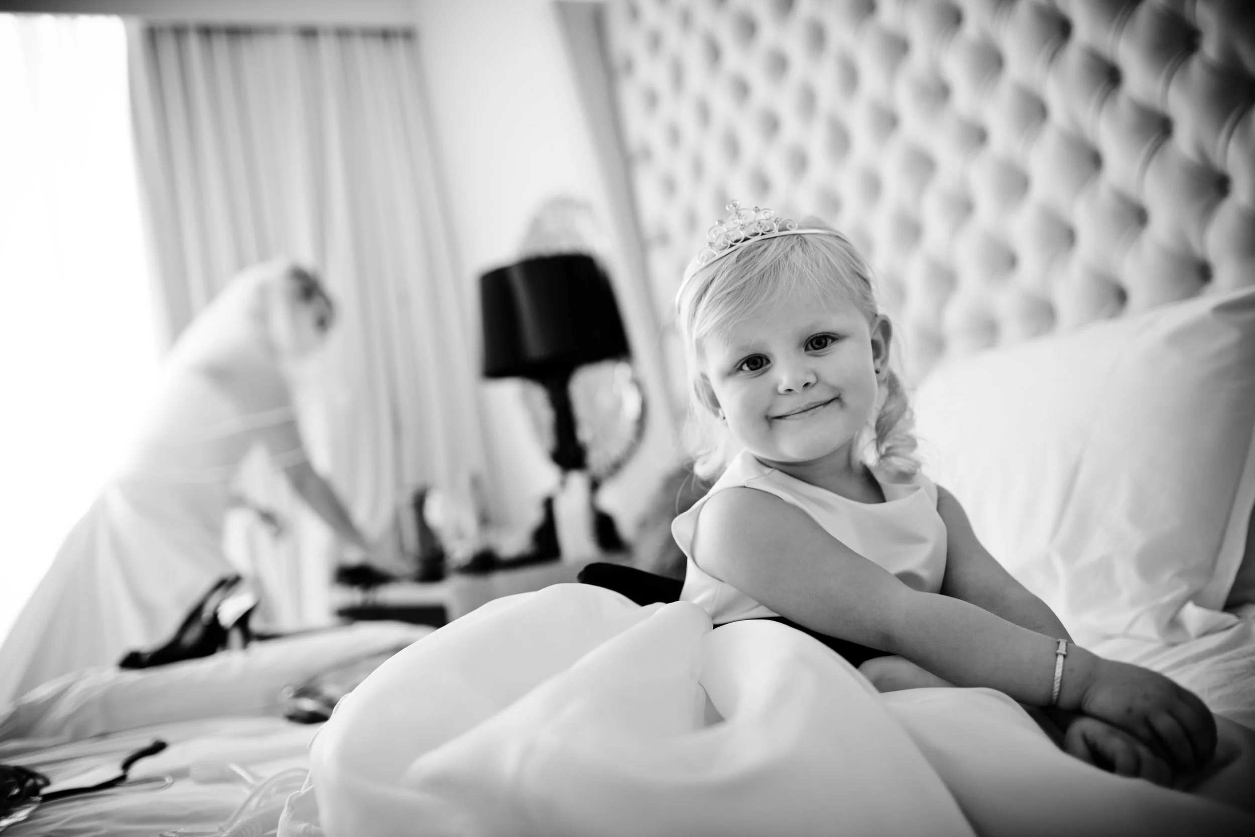 A young girl with blonde hair wearing a tiara and smiling, sitting on a bed in a room with tufted headboard and curtains, in black and white.