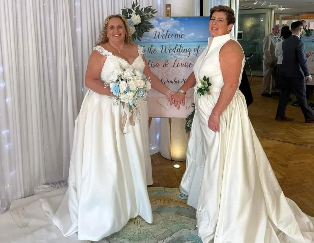 Two women in white wedding dresses holding hands and smiling at a wedding reception, with a welcome sign and other guests in the background.