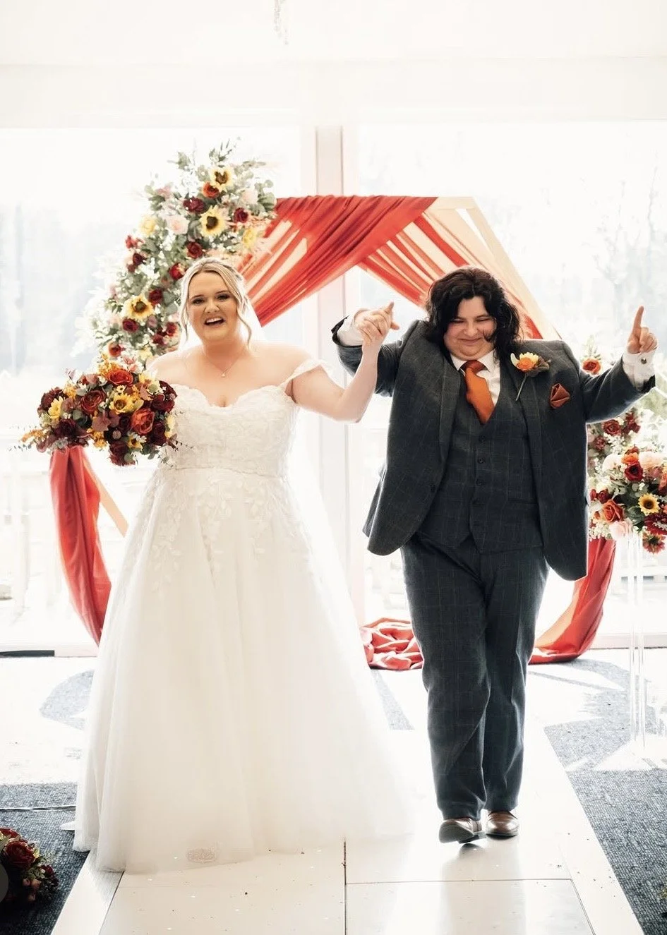A bride and groom walking down the aisle at their wedding ceremony, holding hands and smiling, with decorated floral arrangements and draped fabric in the background.