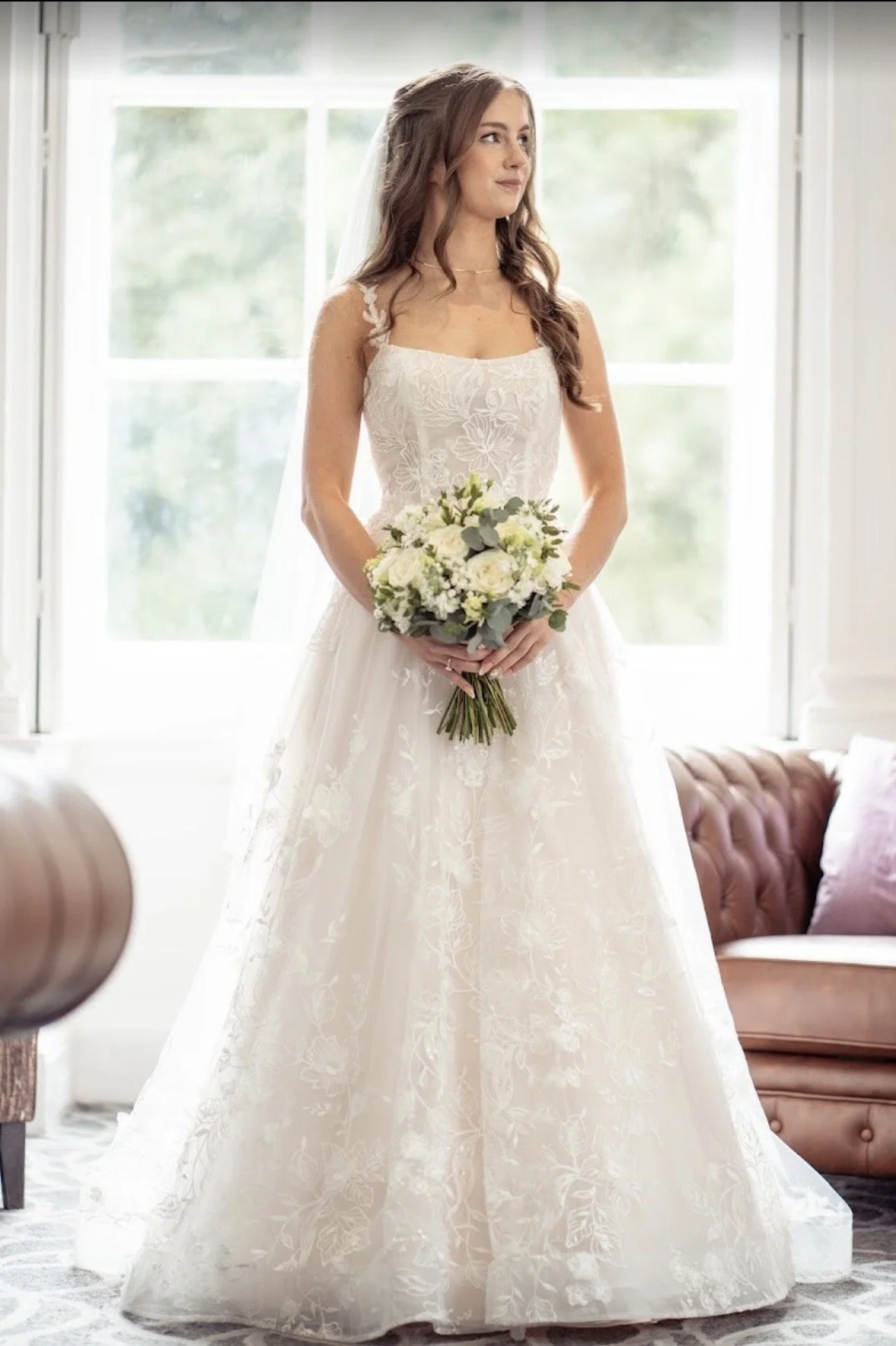 A bride in a white wedding dress holding a bouquet of flowers standing by a window indoors.
