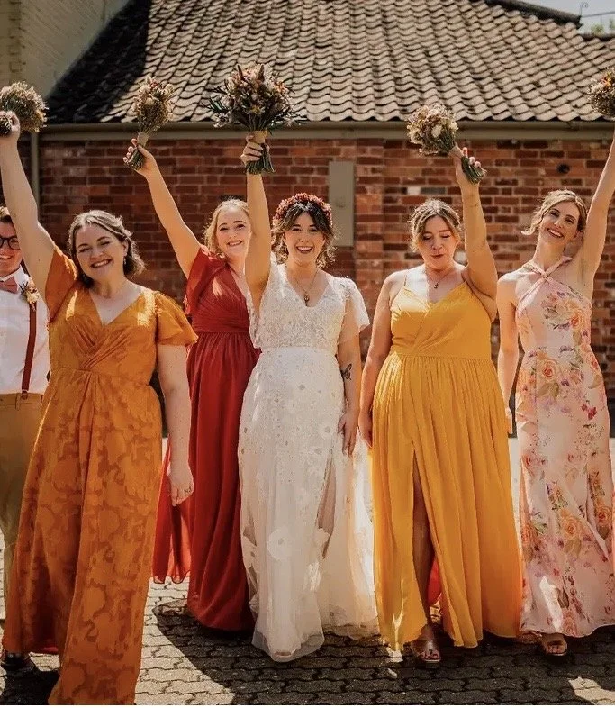 Group of women and one man at a wedding, with women holding bouquets, walking on cobblestone street in front of a brick building, dressed in colorful dresses, celebrating outdoors.