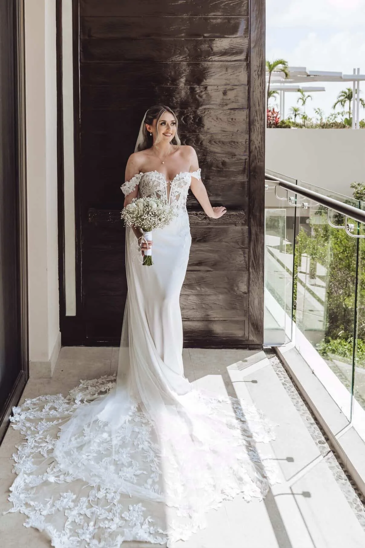 Bride in a lace-trimmed wedding gown holding a bouquet standing on a balcony with a wood-paneled wall behind her and a view of trees and sky outside.