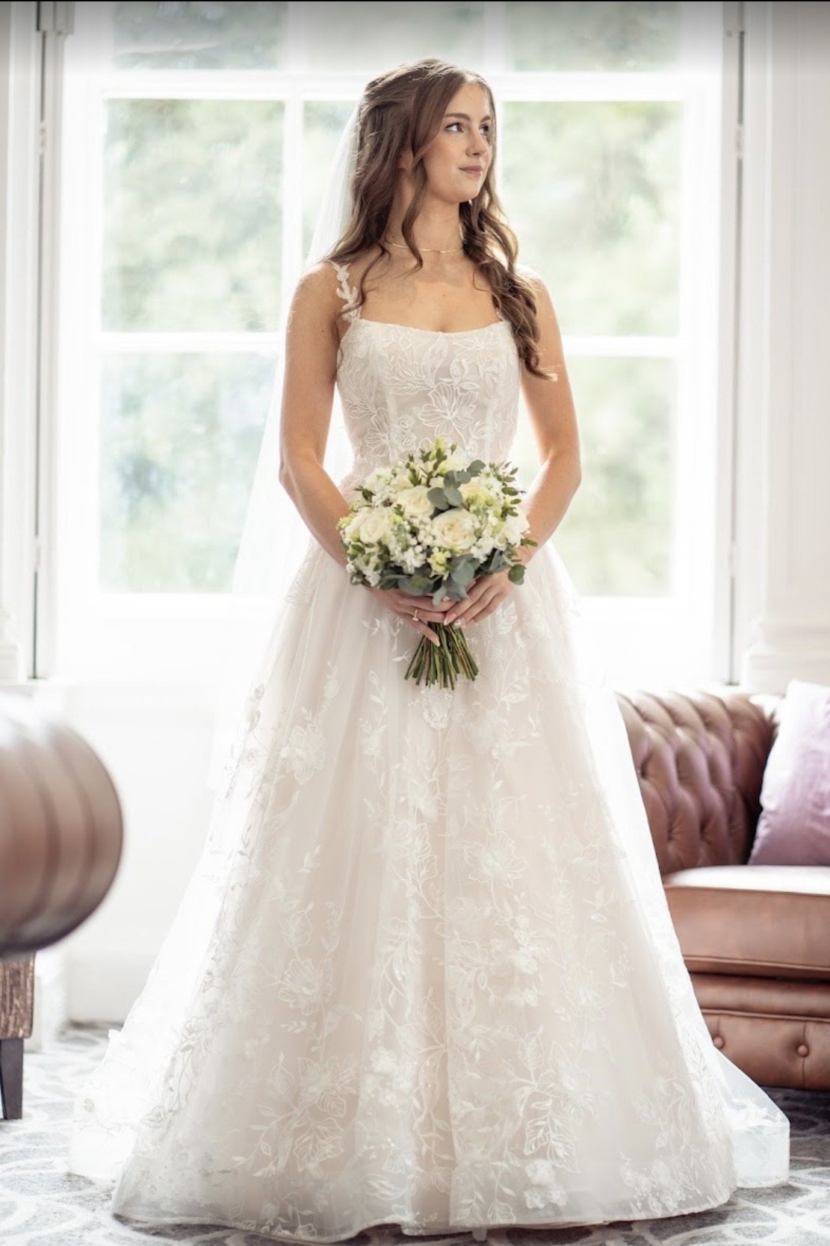A bride in a white lace wedding dress holding a bouquet of flowers inside a bright room in front of large windows.