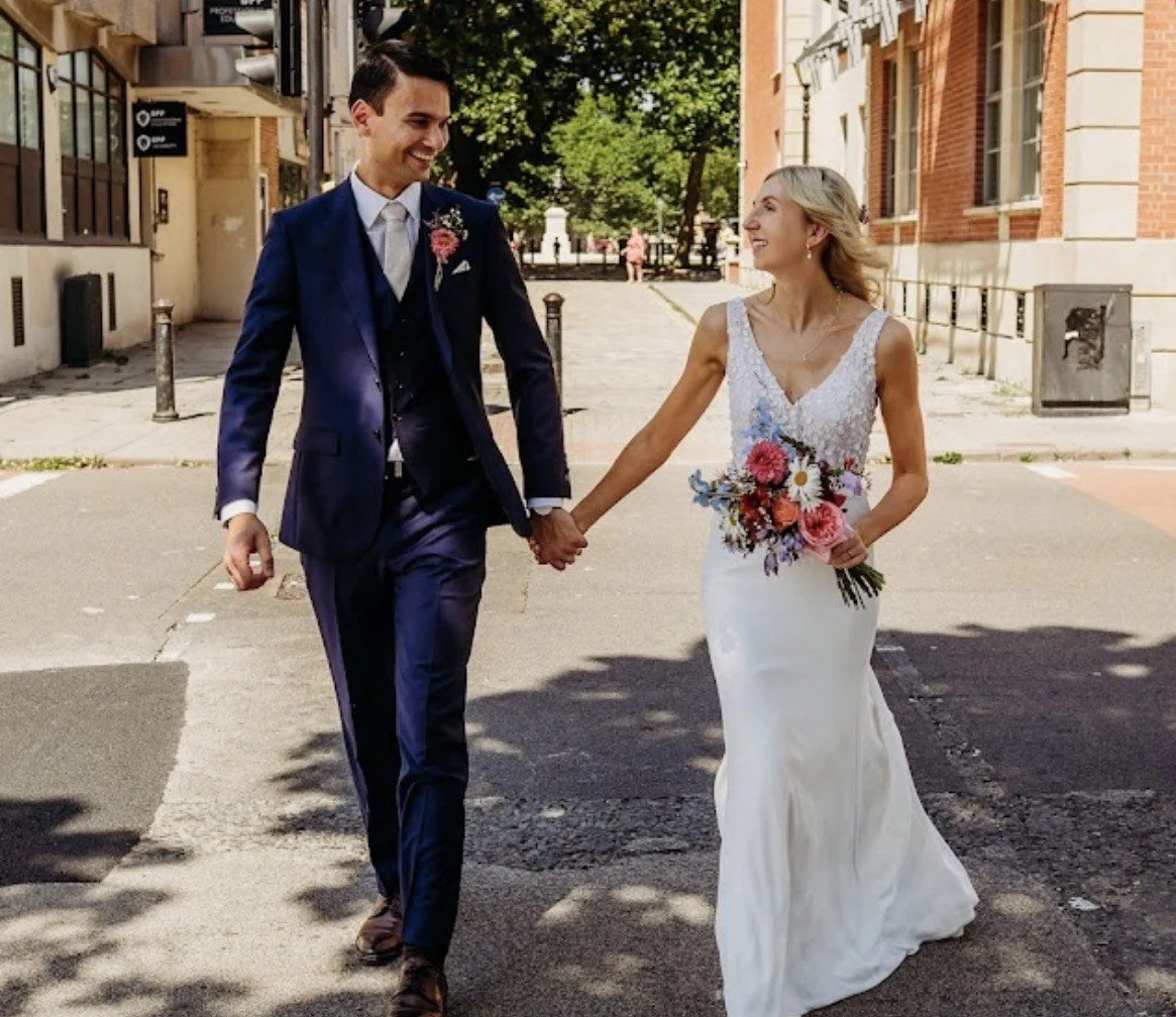 A bride and groom holding hands and smiling during their wedding on a city street. The bride is wearing a white wedding dress and holding a colorful bouquet, while the groom is dressed in a navy blue suit and tie.