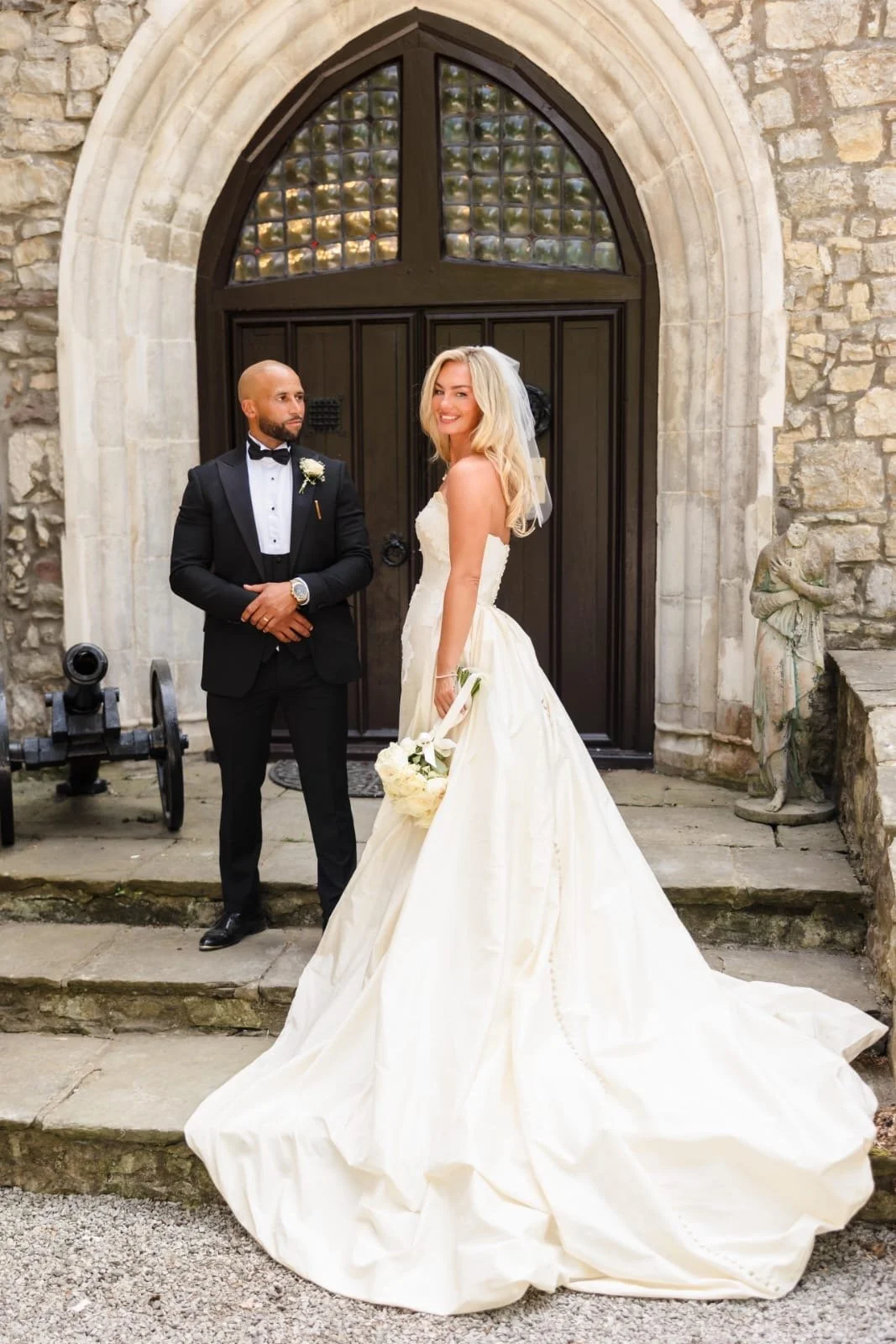 A bride with blonde hair in a white wedding gown holding a bouquet, smiling, standing in front of a dark wooden arched door with an older man in a black tuxedo beside her, outside a stone building. Barry, Vale of Glamorgan. Thread Studio Barry.