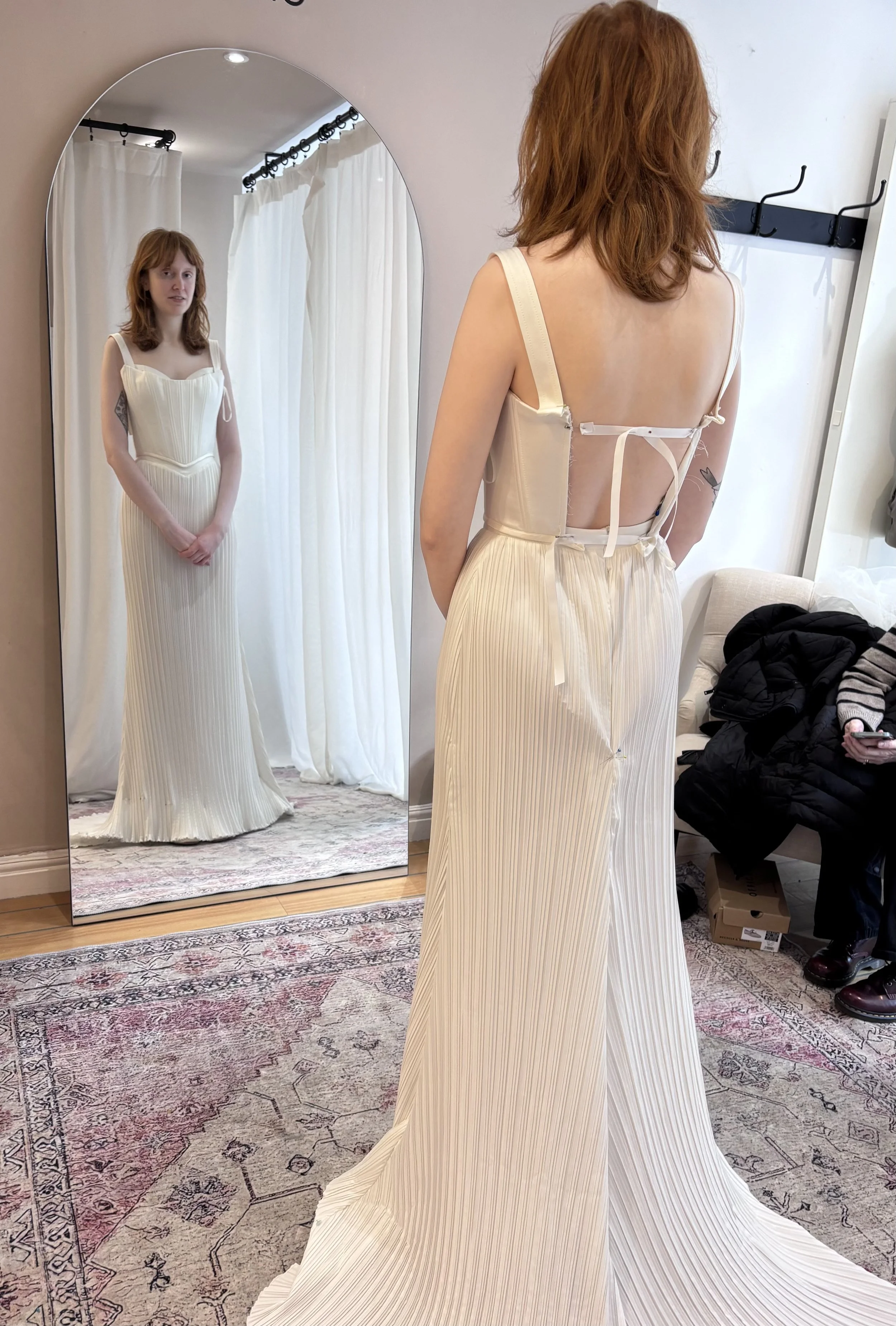 A woman with red hair trying on a cream-colored pleated dress with an open back inside a fitting room. She is standing in front of a large mirror, looking at her reflection. The room has white curtains, a patterned rug, and a coat rack on the wall.