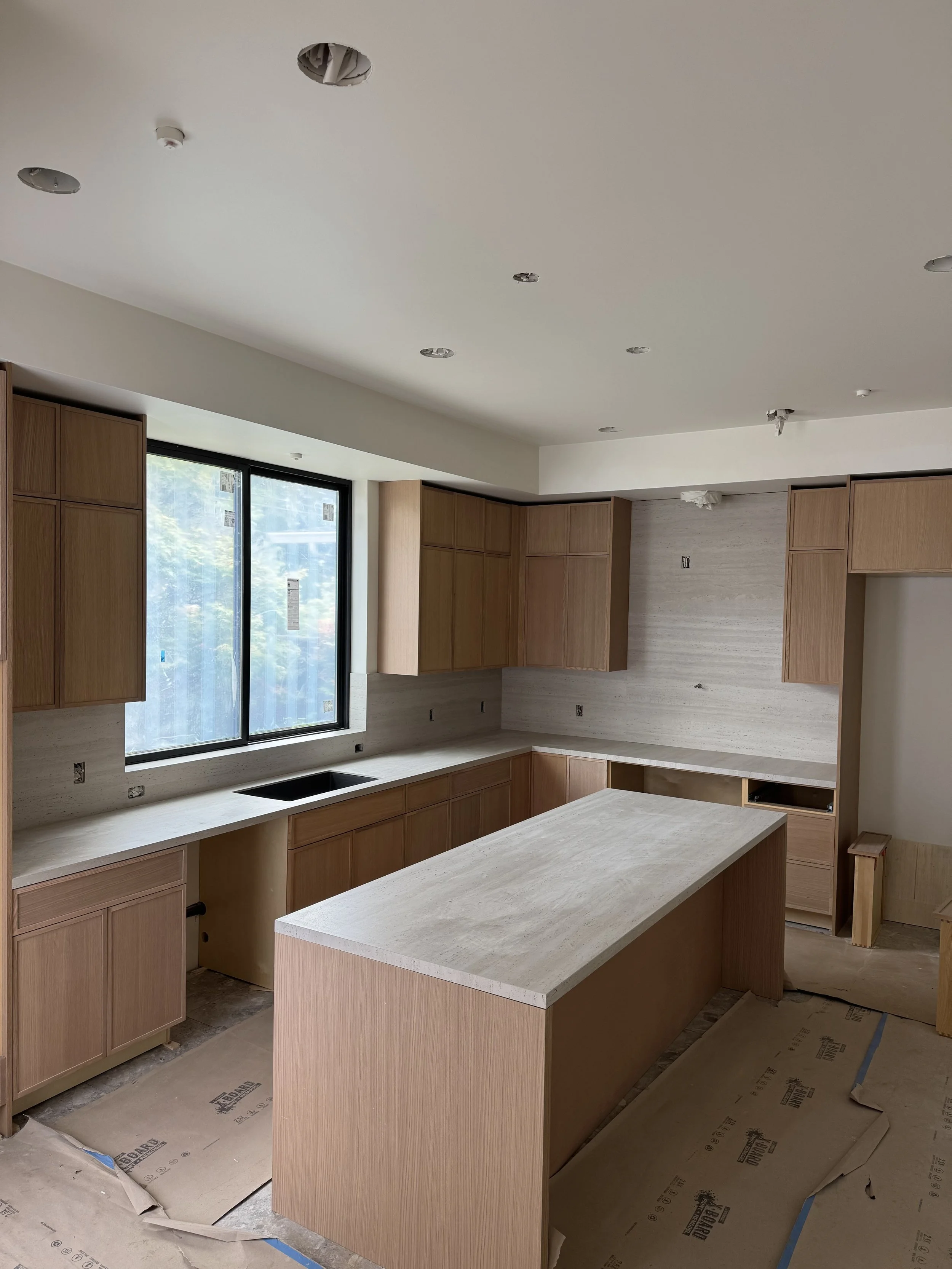 Kitchen under construction with unfinished cabinets, a large window, and a kitchen island, with protective paper on the floor.