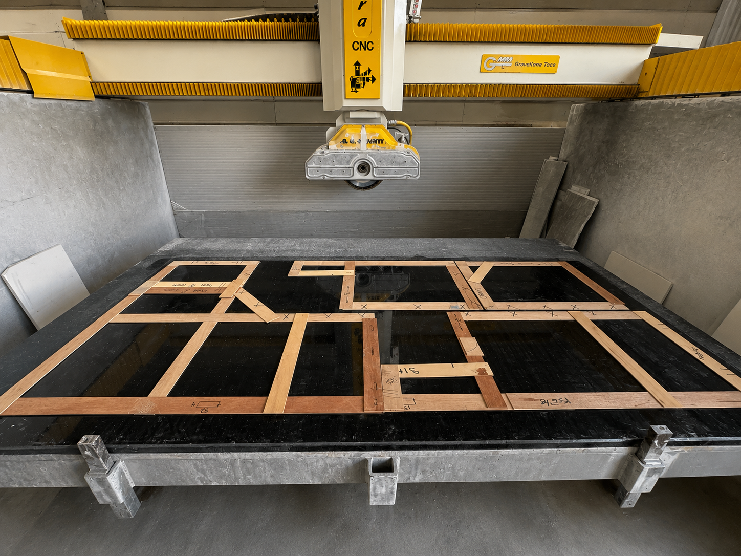 A CNC machine cutting a wooden framework on a black granite surface, with yellow safety features and various pieces of stone and wood around.