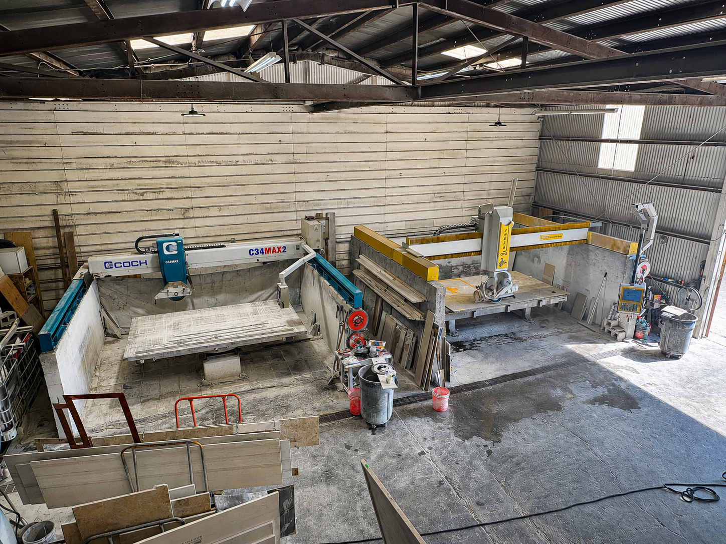 An industrial workshop with two large CNC cutting machines, one labeled 'COACH C34MAX' and the other 'Putsch Meniconi'. There are various tools, materials, and a trash bin around, with a concrete floor and a corrugated metal roof.