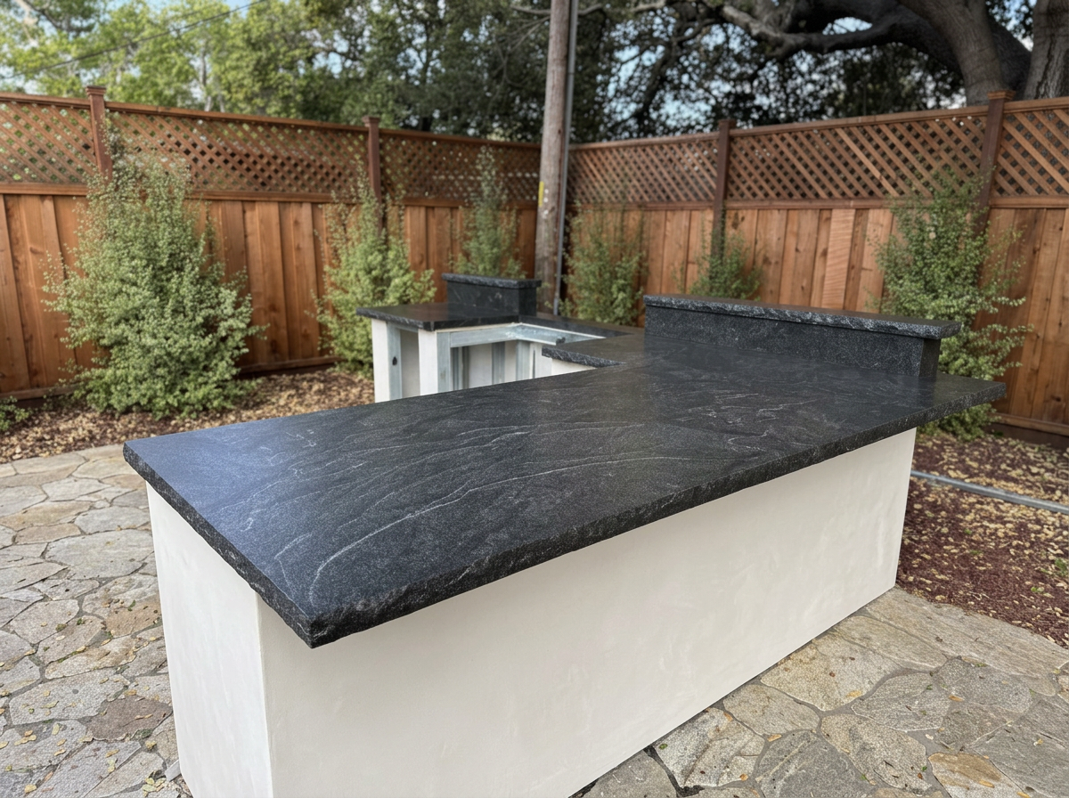 Outdoor kitchen with black granite countertop and white base, with a wooden fence and trees in the background.