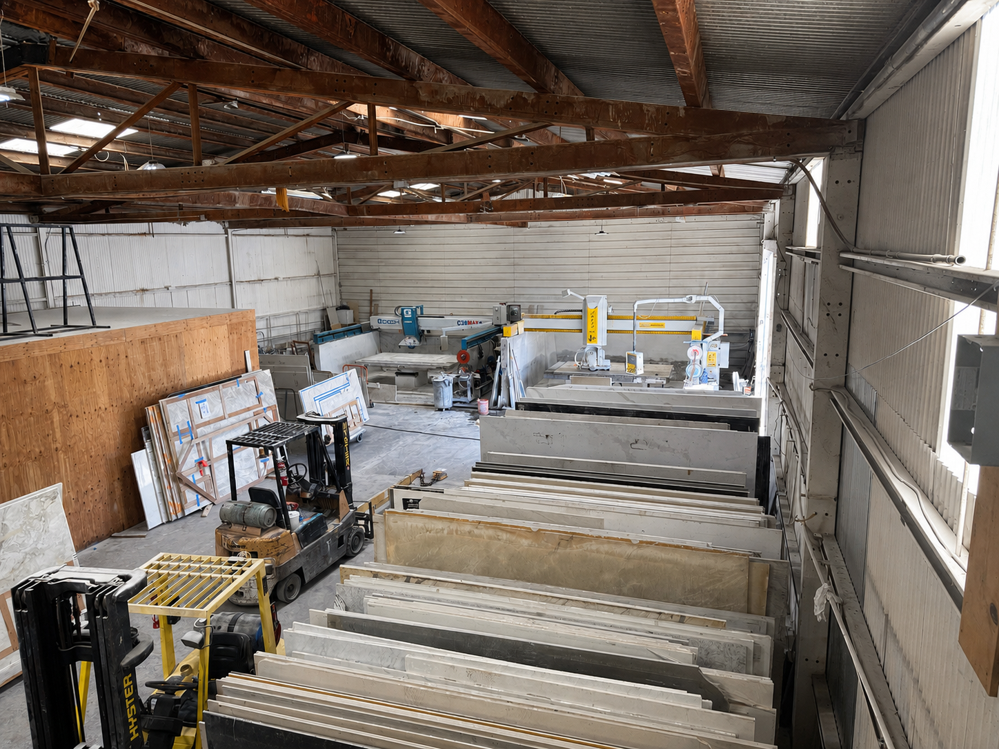 Inside a warehouse or workshop with large stone or marble slabs stored vertically and machinery for cutting or processing stone in the background.