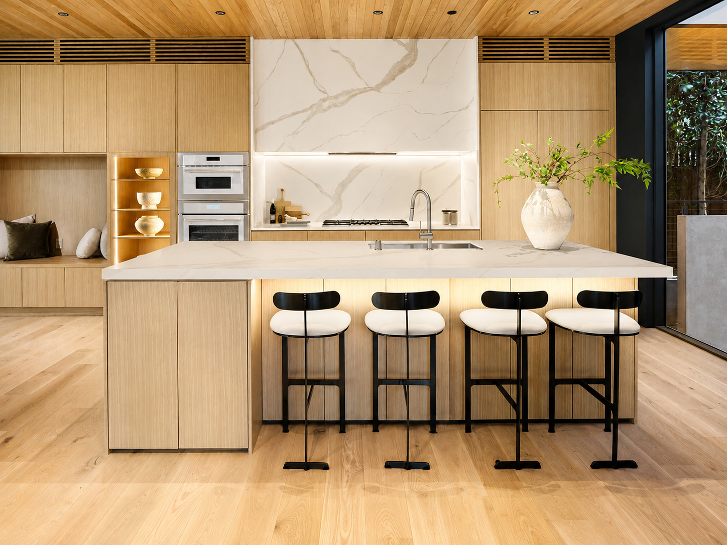 Modern Kitchen with wooden cabinets, marble backsplash, four black and white bar stools at a marble island, decorative vase with green branches, built-in oven and microwave, and large window with outdoor view.