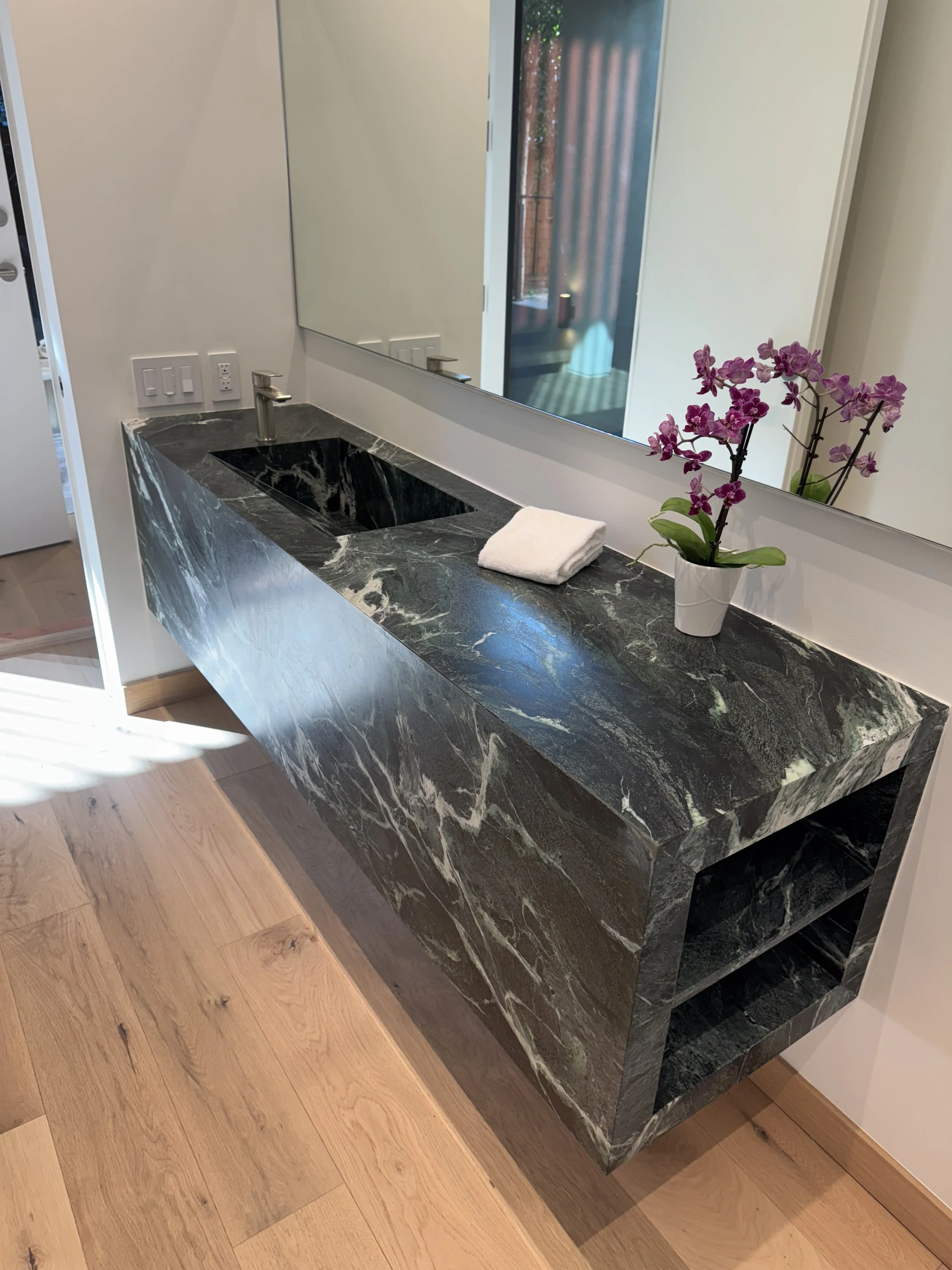 A bathroom vanity with a dark green and white marble countertop, a rectangular undermount sink, a silver faucet, a folded white towel, a potted purple orchid, and a large mirror reflecting an outdoor area with a brick wall and a patio.