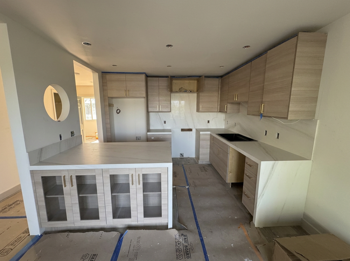 Kitchen under construction with light wood cabinets, marble countertops, and a black sink, with packaging and protective coverings on the floor.