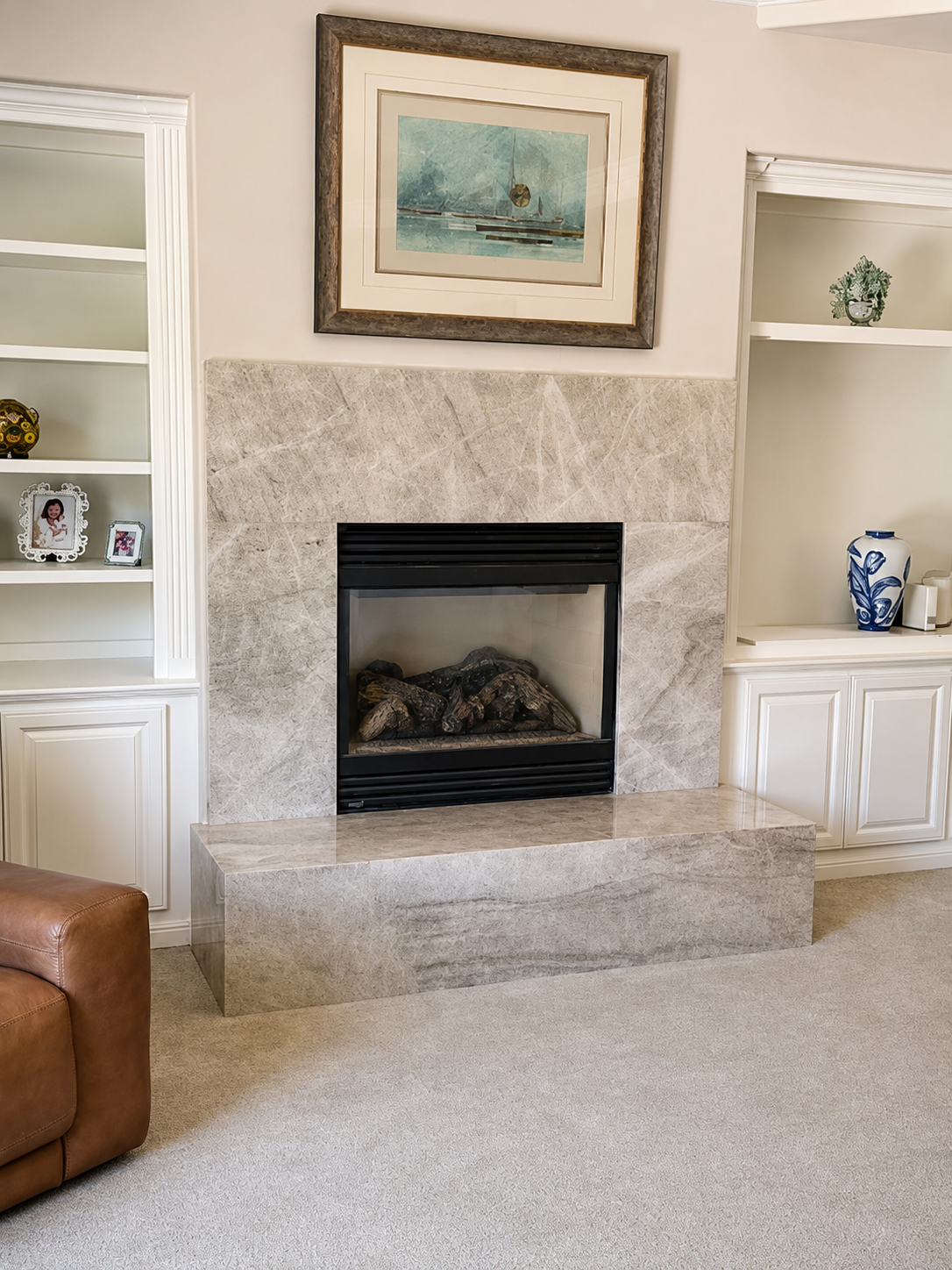 Living room with a modern fireplace framed by beige stone, flanked by white built-in shelves with decorative items, a framed nautical painting above, a brown leather armchair in the lower left corner, and beige carpet flooring.