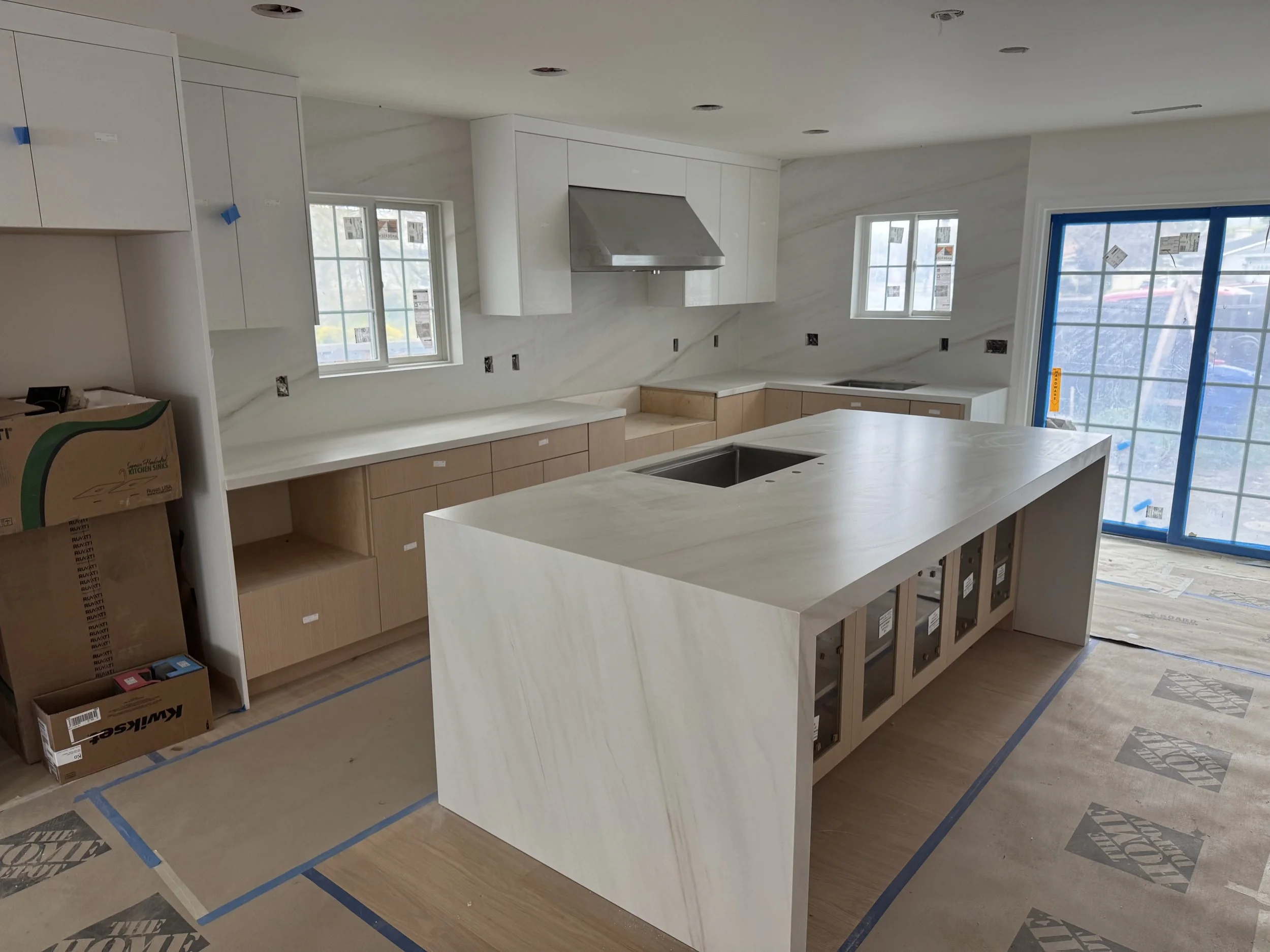 Interior of a kitchen under construction with white cabinets, containing no countertops, an island, and multiple windows, with protective tape on the floor.