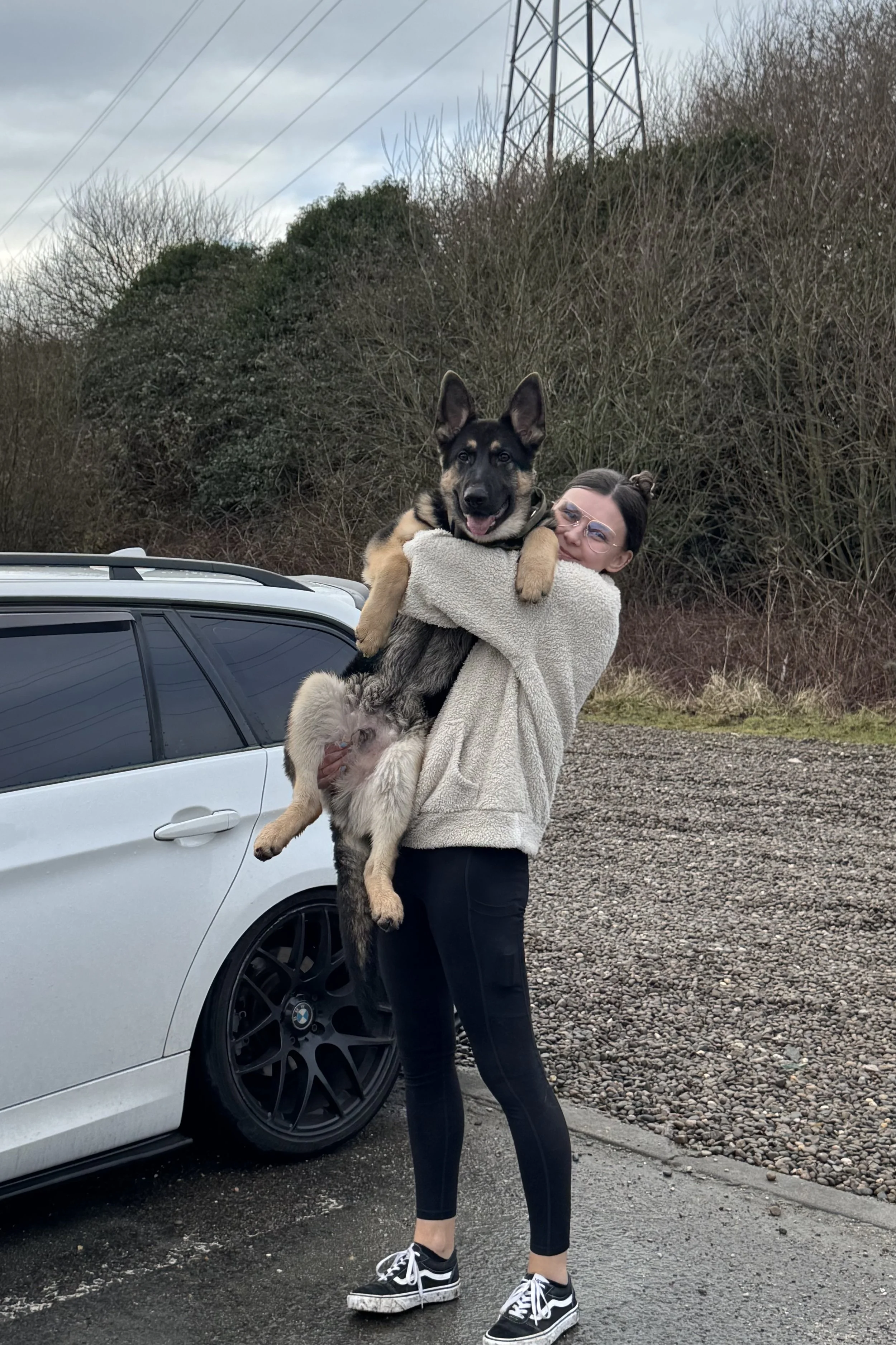 A woman holding a large German Shepherd dog next to a white car parked on a gravel lot. The woman has dark hair tied in a bun, glasses, and is wearing a beige fleece jacket, black pants, and black and white sneakers. In the background, there are trees and power lines under a cloudy sky.