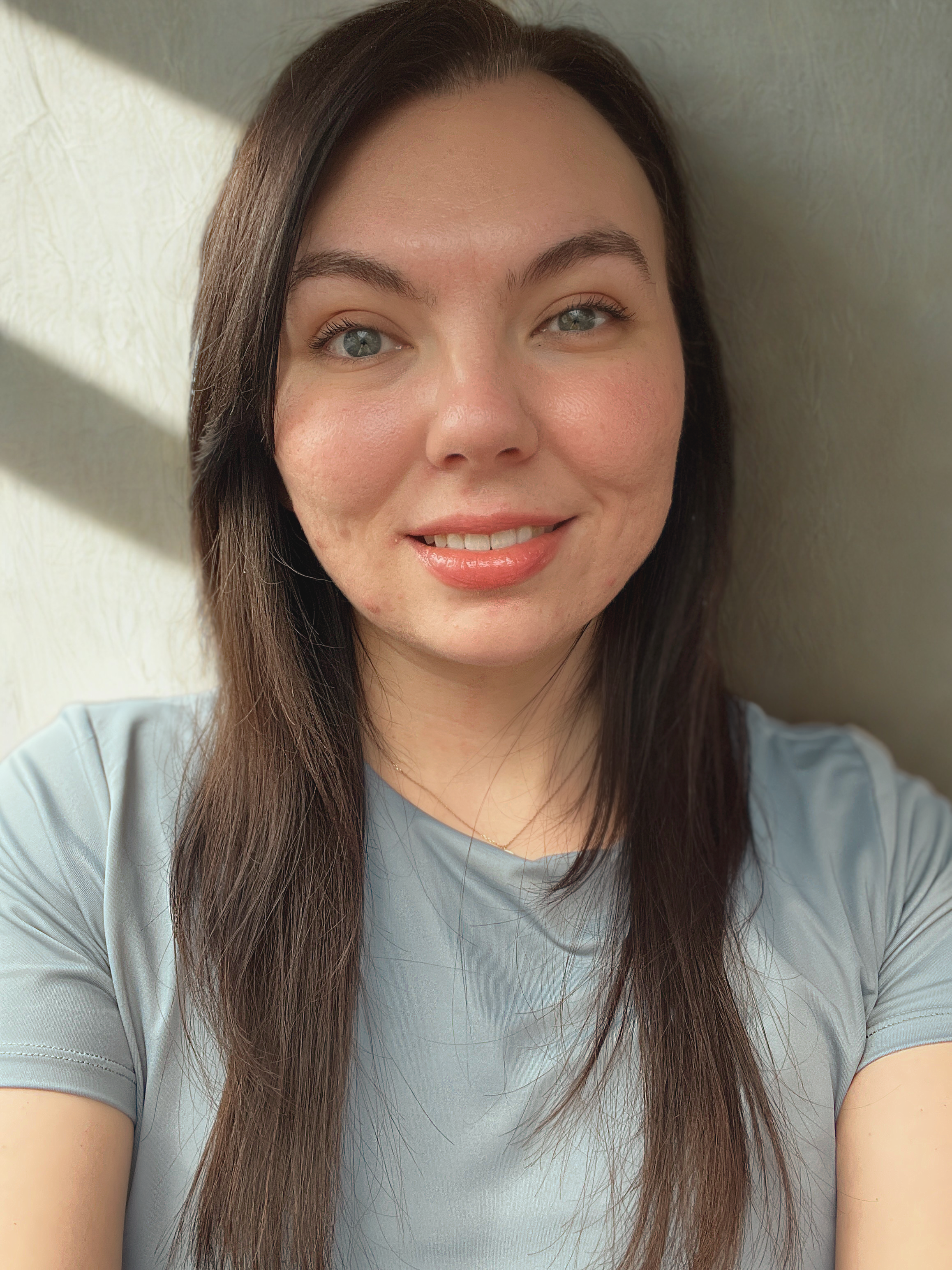 Close-up of a woman with long brown hair, light skin, and blue eyes, smiling while wearing a light gray shirt.