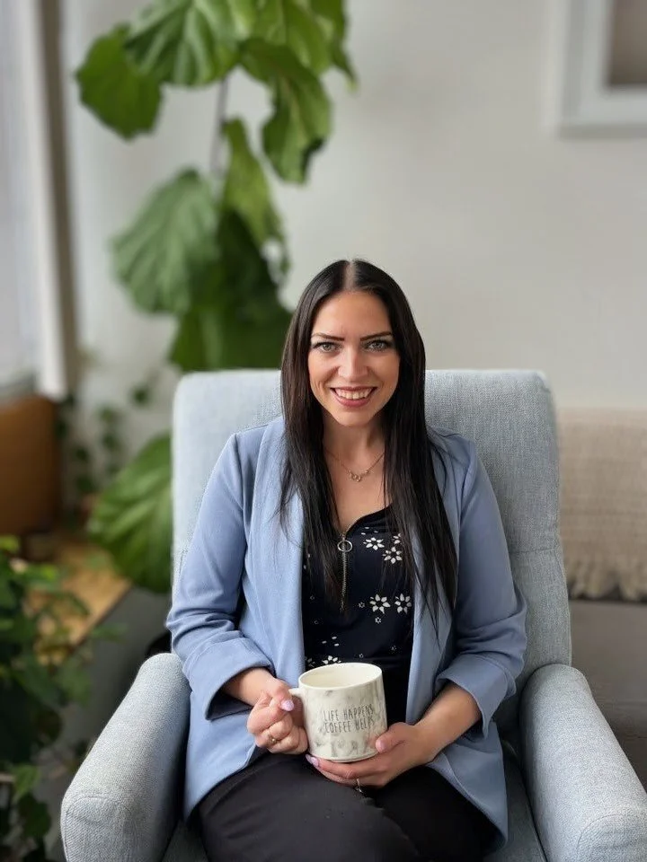 A woman with long dark hair and a blue blazer sitting on a light-colored armchair, holding a coffee mug that says 'Life happens, coffee helps,' in a well-lit room with green plants in the background.