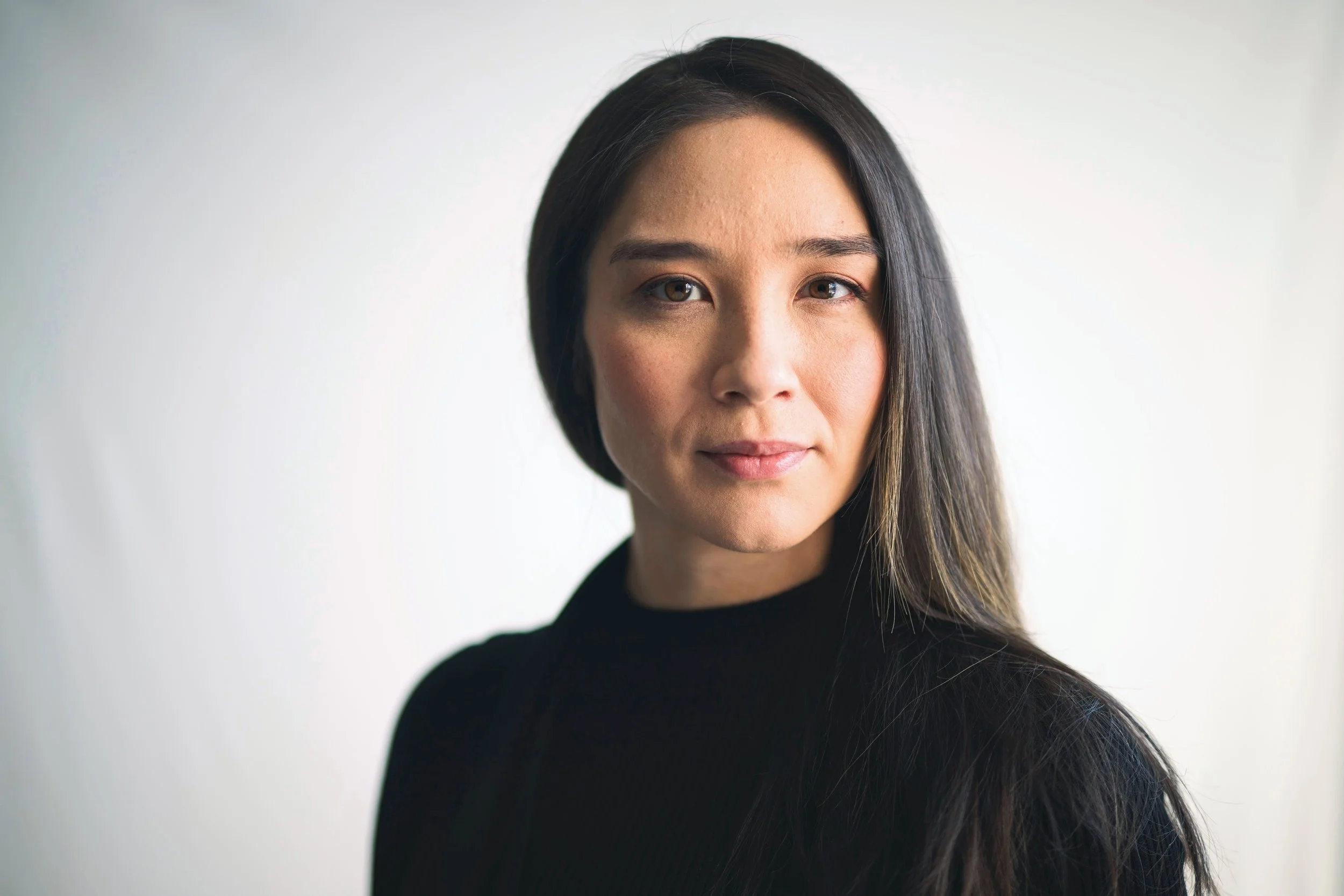 A woman with long dark hair wearing a black top, looking at the camera with a neutral expression, against a plain light background.