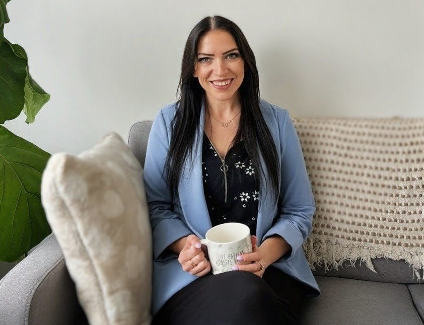 A woman with long black hair, wearing a blue blazer over a black shirt with white patterns, sitting on a gray sofa, holding a white mug, smiling at the camera.