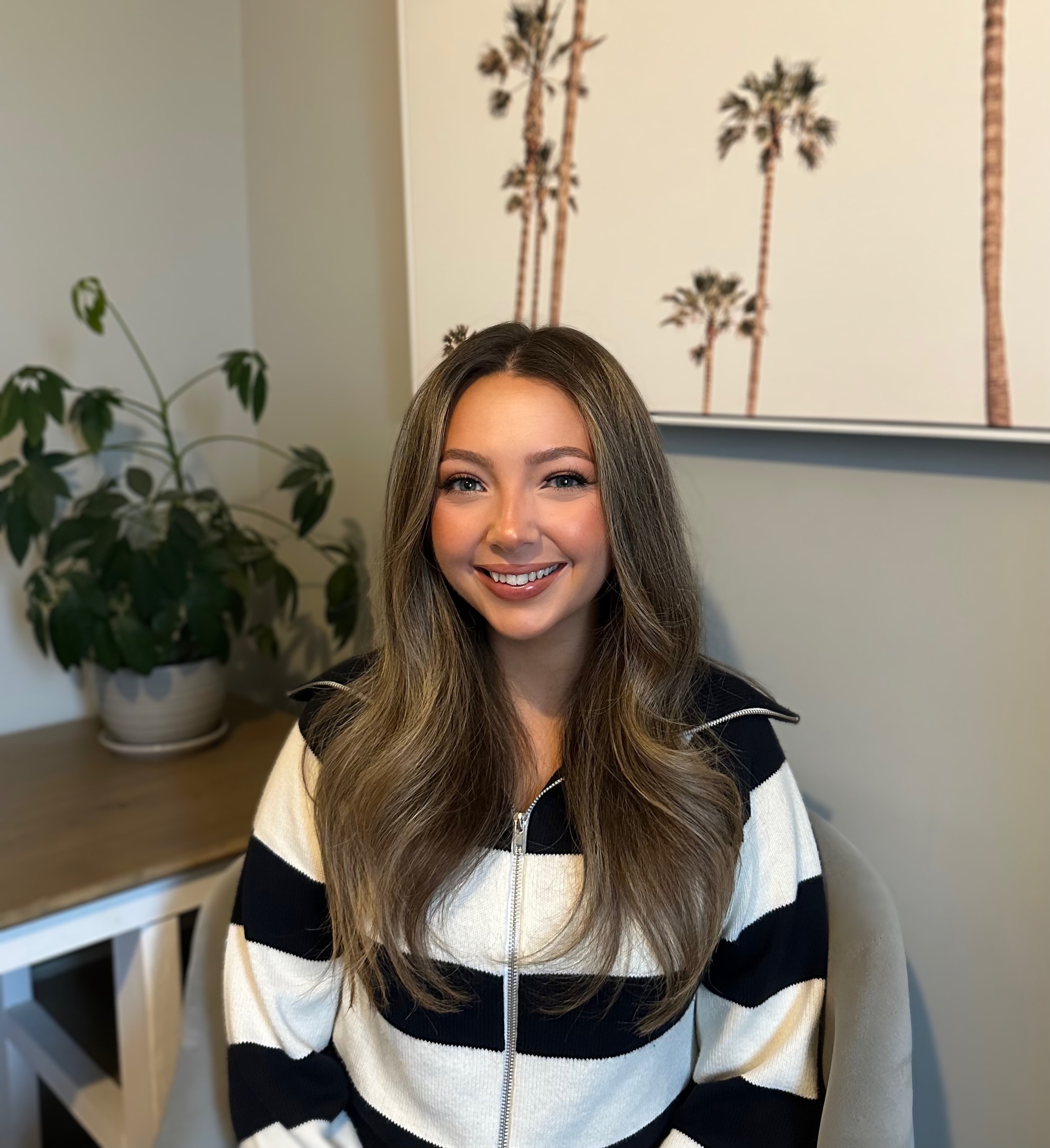 Smiling young woman with long brown hair sitting indoors, with a potted plant and a framed art piece of palm trees in the background.