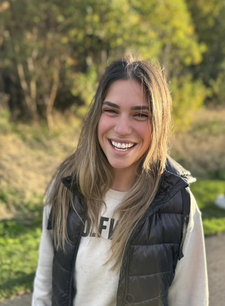 A young woman with long, light brown hair, smiling outdoors in sunlight, wearing a black puffer vest over a light-colored sweatshirt, with trees and greenery in the background.