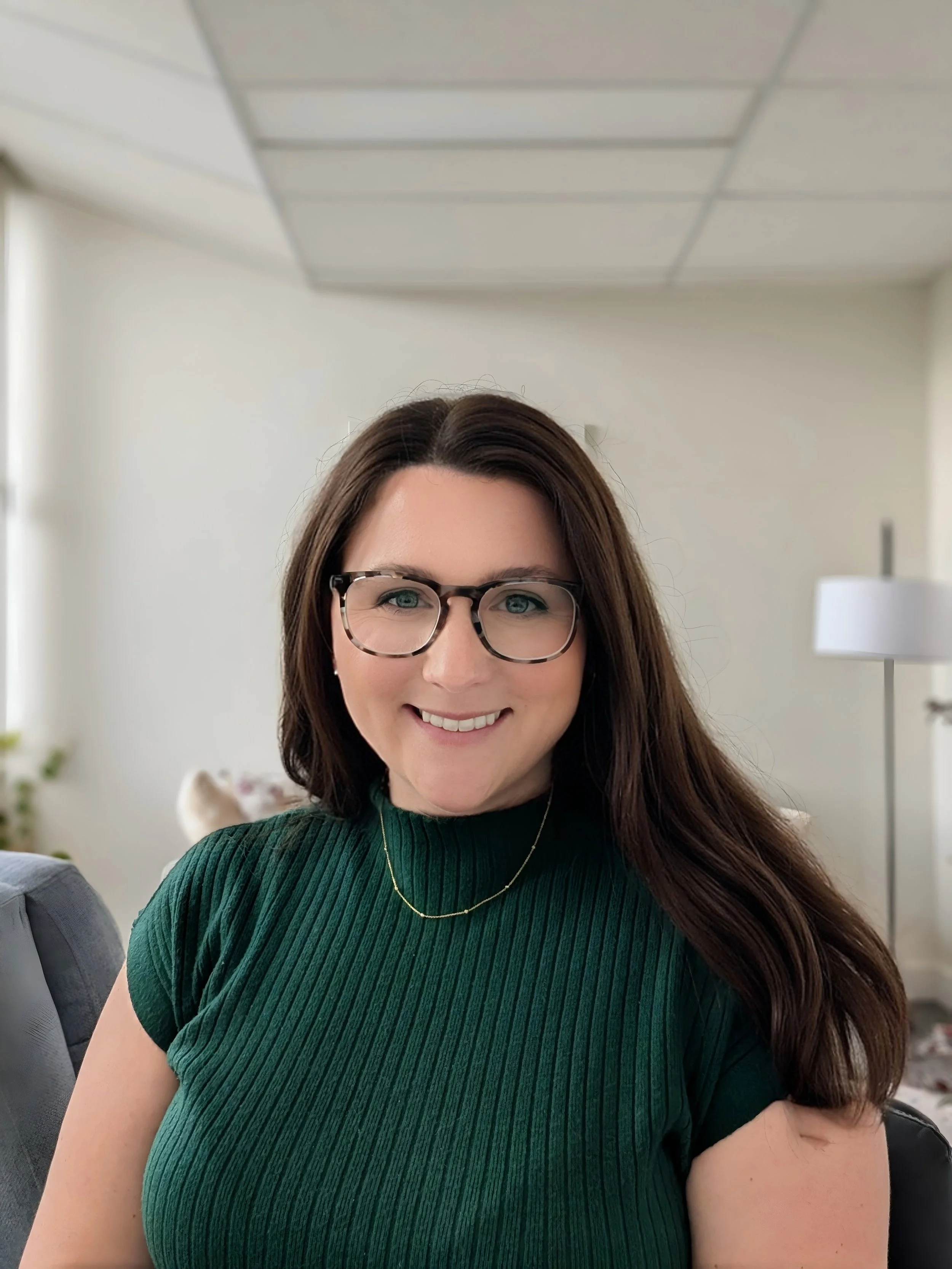 A woman with brown hair, glasses, wearing a green ribbed top and a gold necklace, smiling in an indoor setting with a lamp and a white wall in the background.