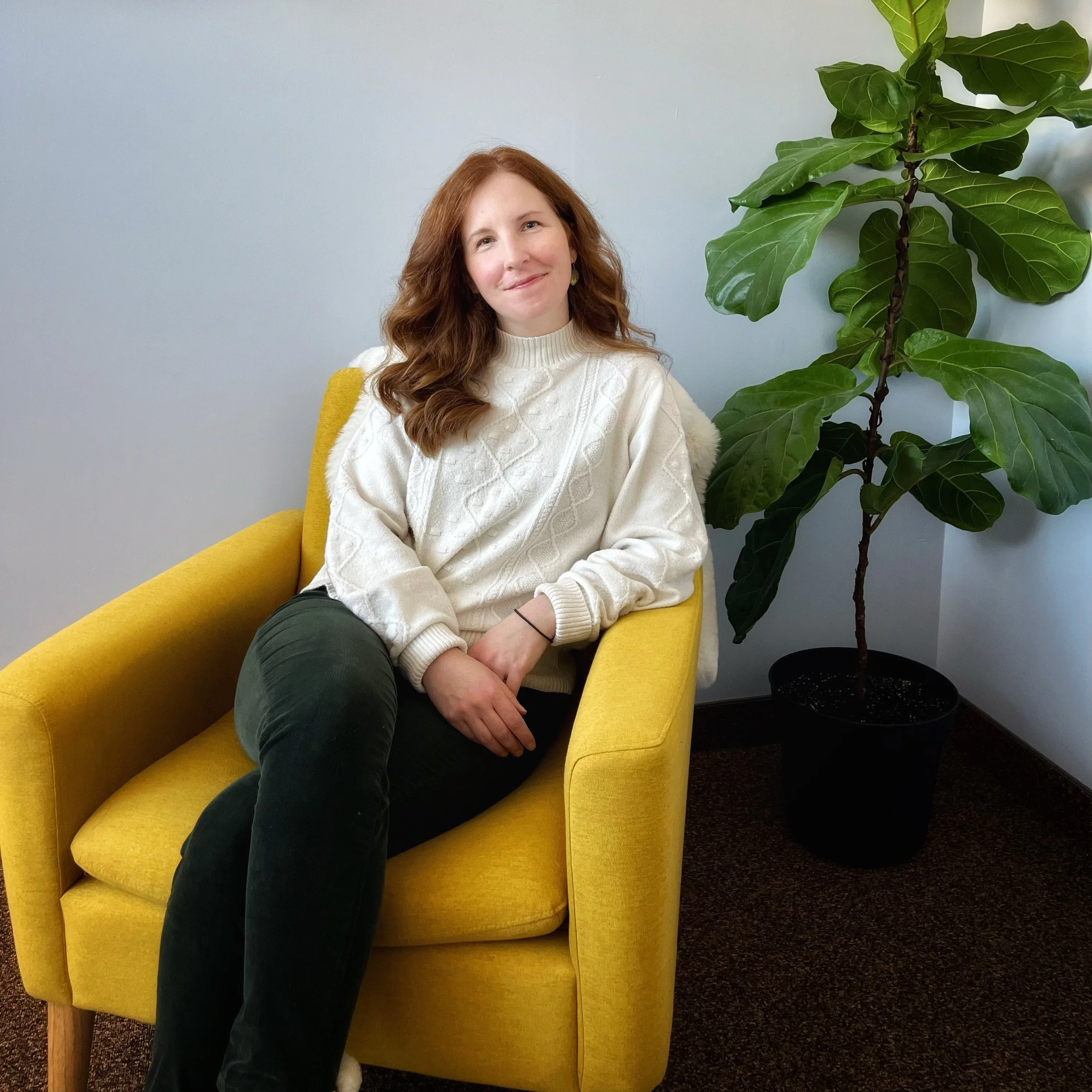 A woman with curly red hair sits on a yellow chair next to a large green plant, against a light gray wall.