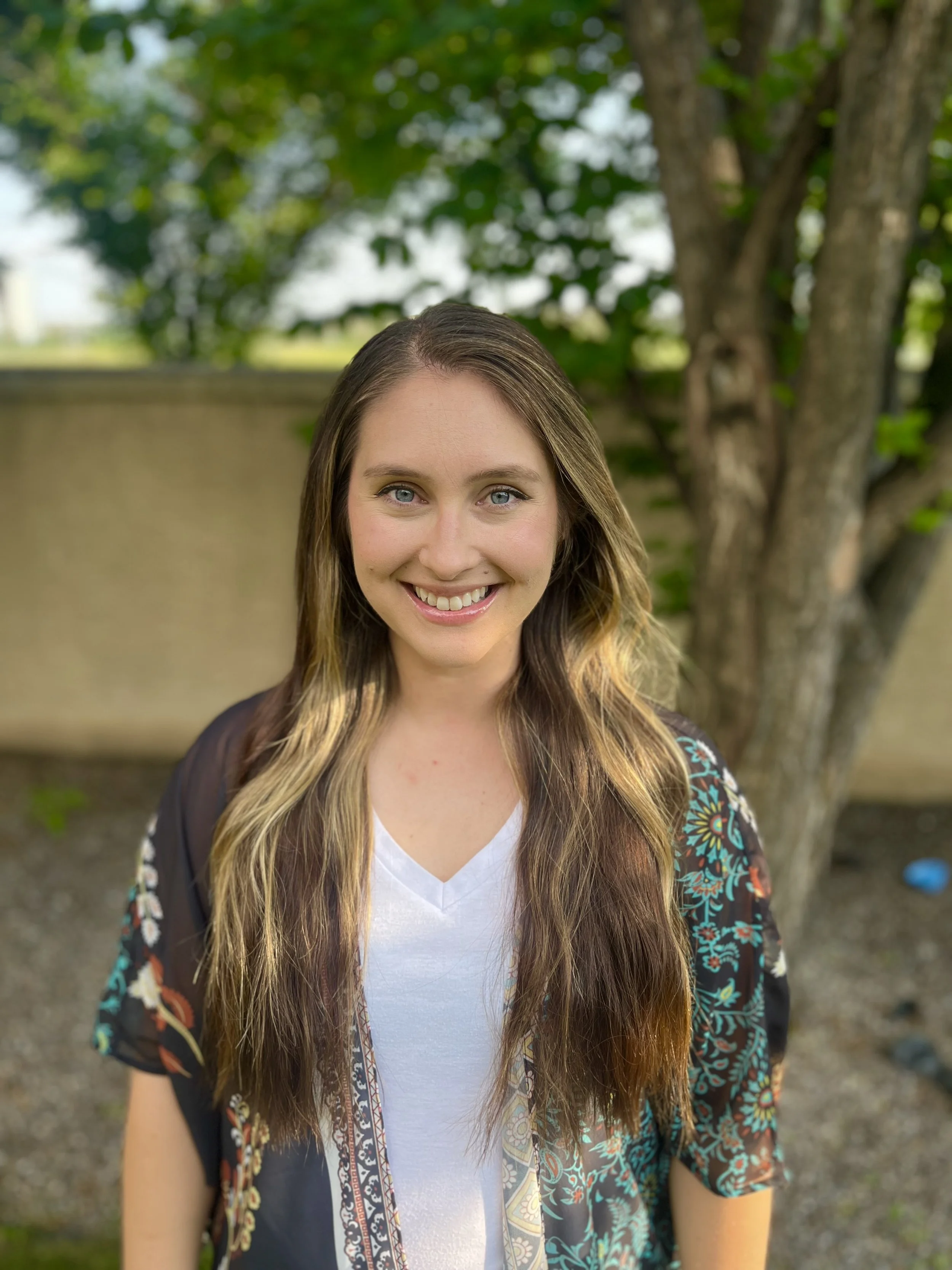 A young woman with long, wavy hair and blue eyes smiling outdoors with a tree and green foliage in the background.