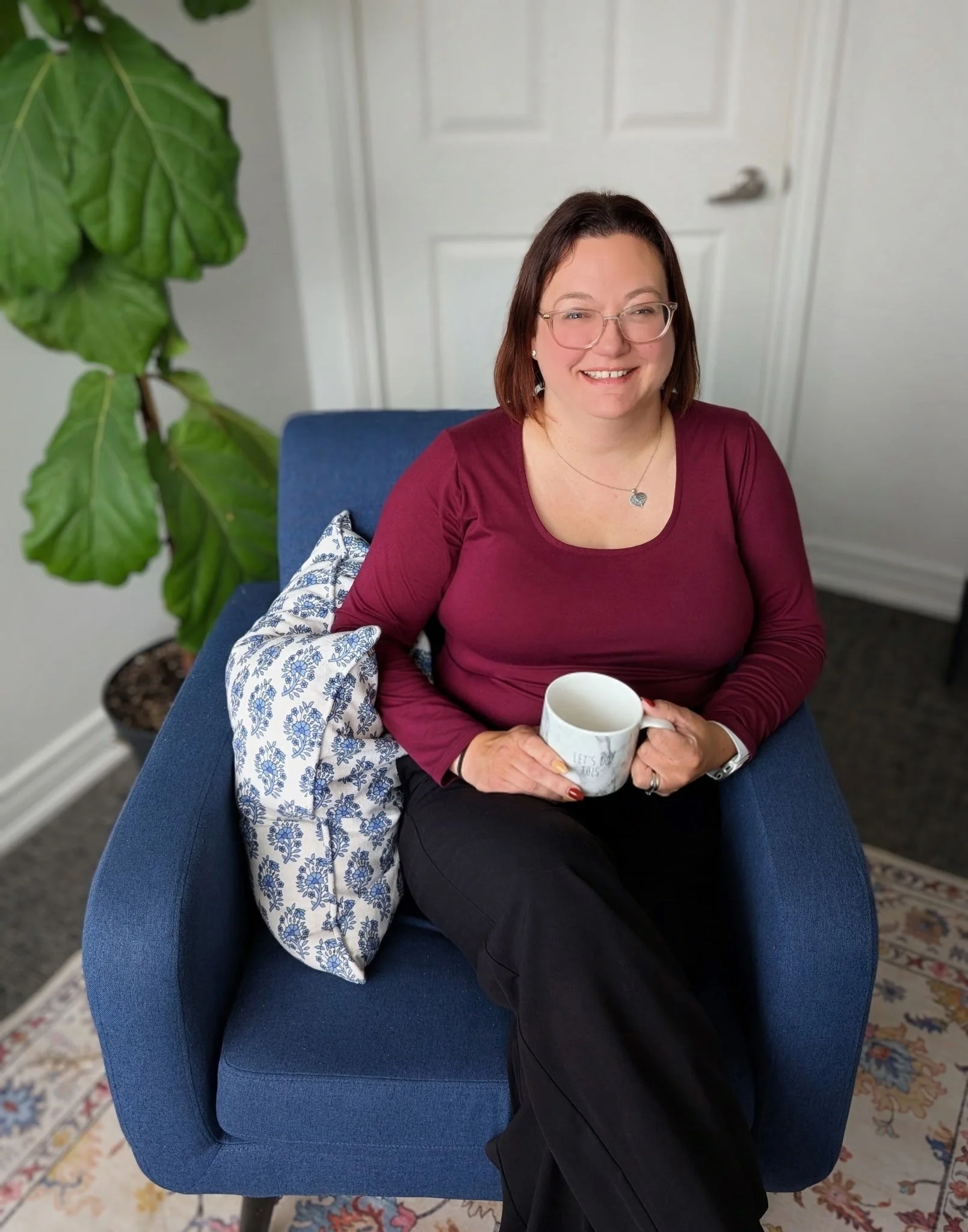 A woman with shoulder-length brown hair, wearing glasses and a burgundy long-sleeve shirt, seated on a blue chair, holding a white mug with a quote, smiling. A large green houseplant is behind her on the left, and a white door is in the background.