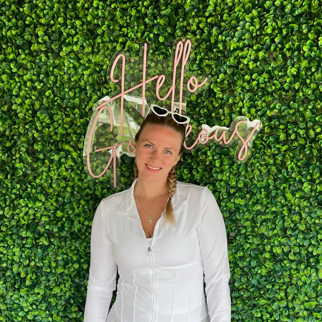 A woman with a braid and sunglasses on her head, smiling, standing in front of a green leafy wall with pink neon sign that says 'Hello Gorgeous'.