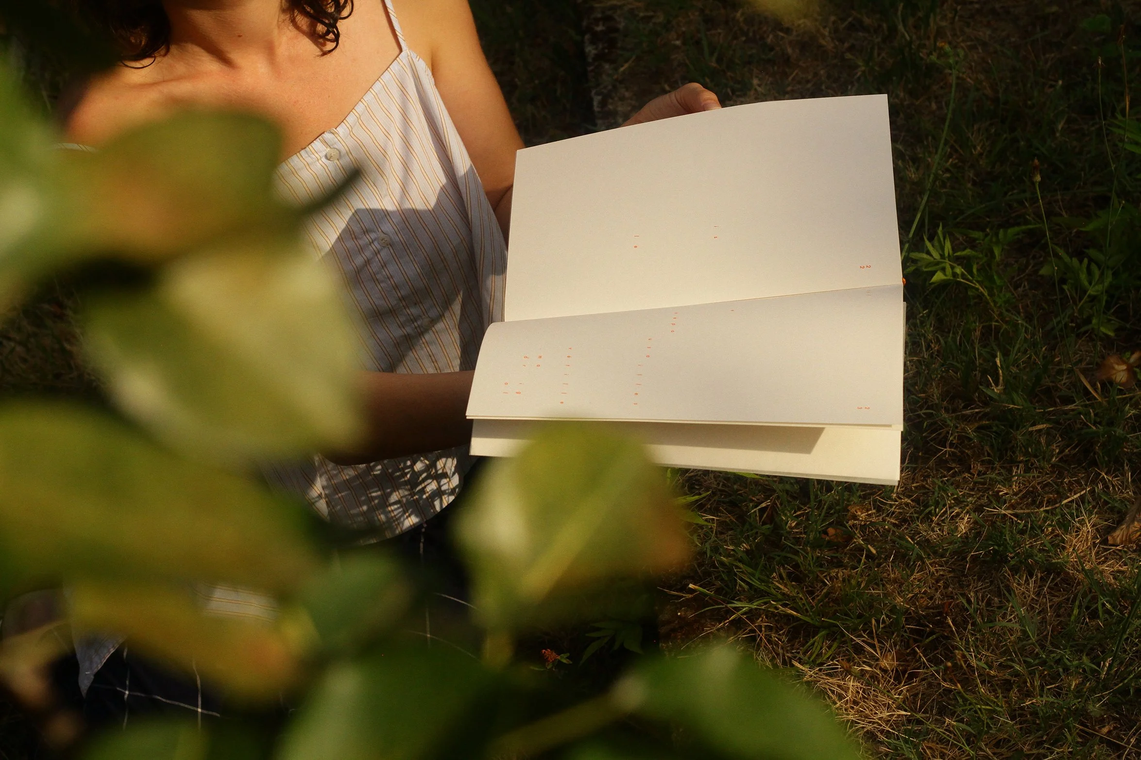 A woman with curly brown hair wearing a striped sleeveless top, holding an open blank book or notebook outdoors surrounded by grass and foliage.