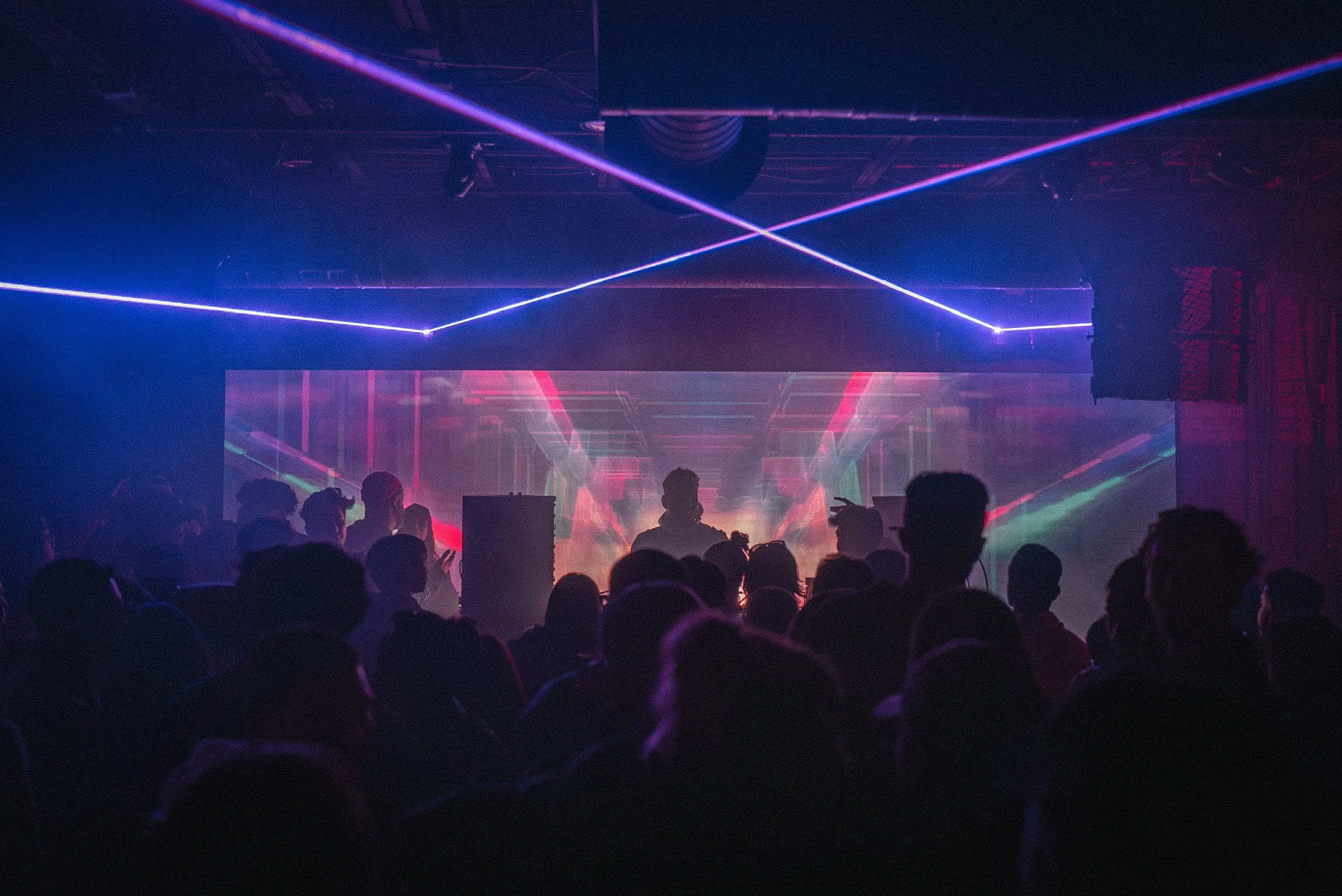 A concert scene with a crowd of people facing a stage illuminated by colorful laser lights and LED screens, creating a vibrant, energetic atmosphere.