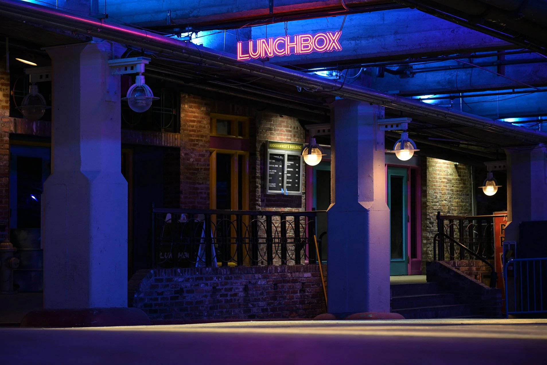 Nighttime exterior of Atlanta club Lunchbox that reads 'LUNCHBOX' overhead. The scene is lit with blue and purple lighting, and there are hanging lamps and a menu board on the wall.