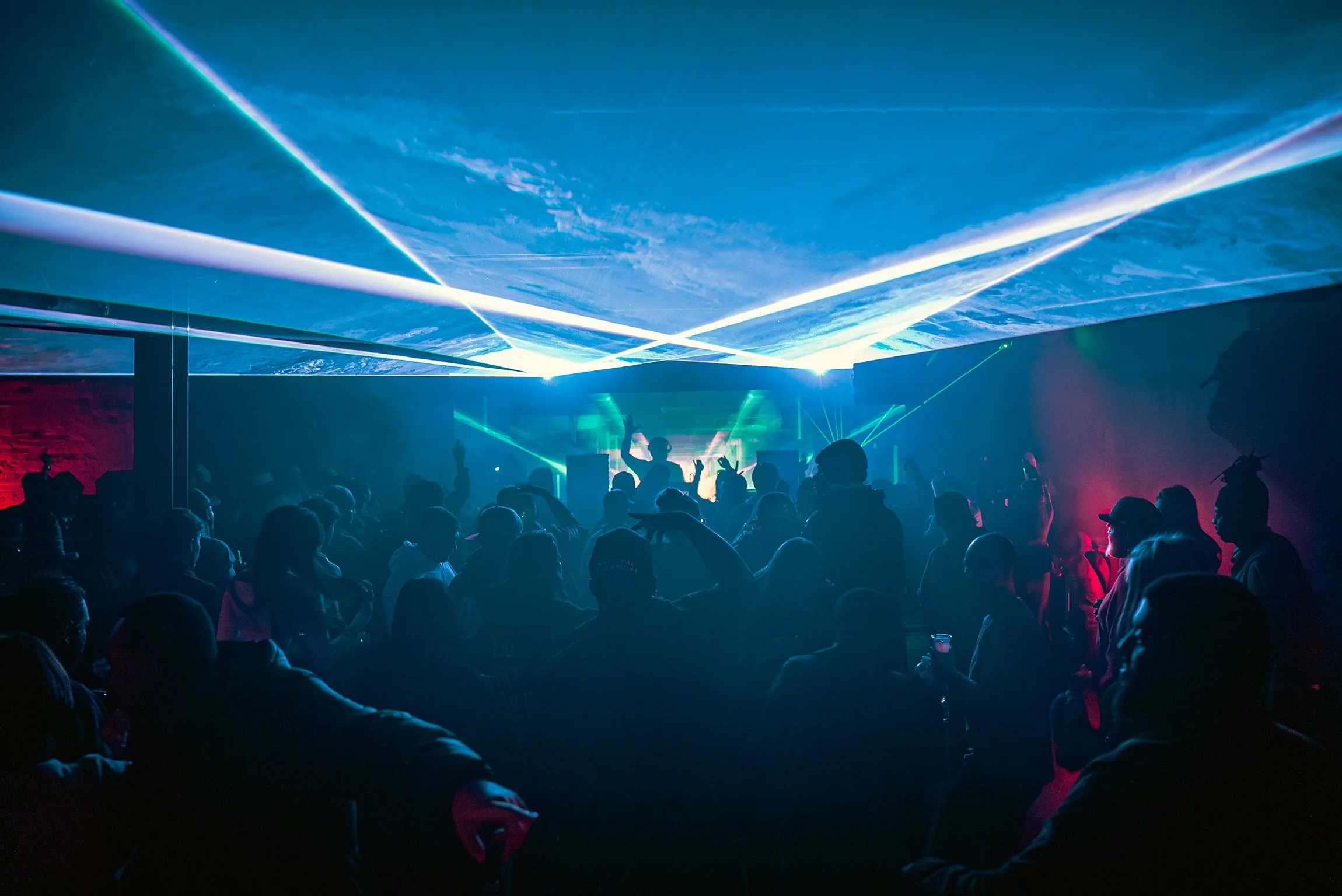 Lunchbox Atlanta with a crowd of people watching a performer on stage. The stage features bright, colorful laser and LED light effects illuminating the space in green, blue, and purple hues.