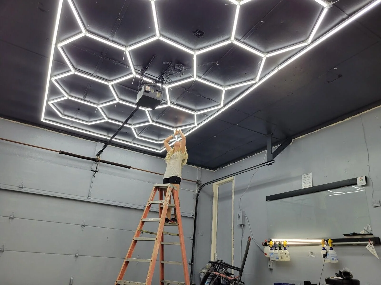 A person standing on a ladder installing or maintaining hexagonal LED light fixtures on a black ceiling in a garage or workshop.