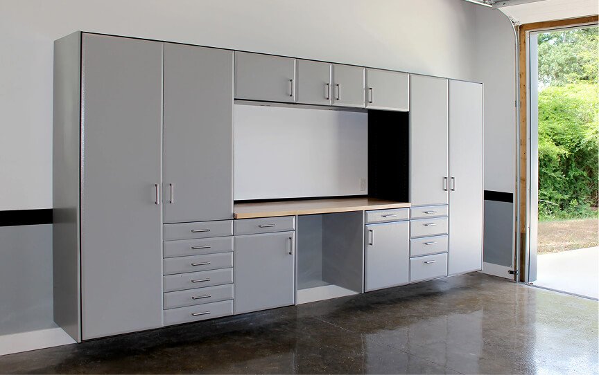 Gray cabinetry with multiple drawers and cabinets, a countertop, and an open space for a TV or monitor, inside a garage or workshop with an open door showing greenery outside.