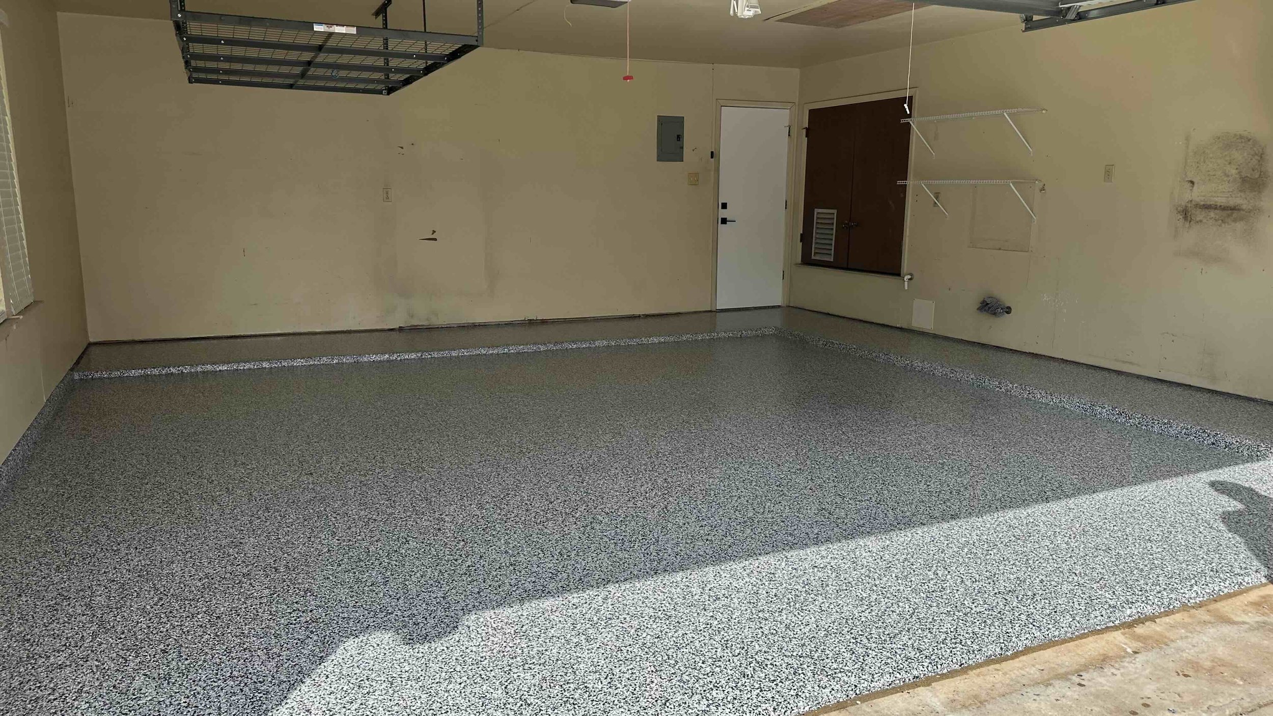 Empty garage with speckled gray epoxy floor, beige walls, and a white door, with hanging shelves and an unfinished section near the floor.
