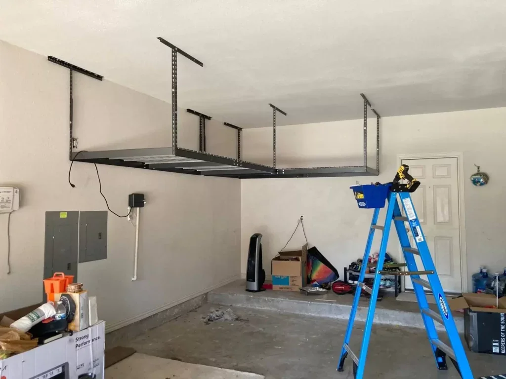 Garage with a steel lift suspended from the ceiling using metal brackets, a blue ladder with tools, boxes, and miscellaneous items on the floor.