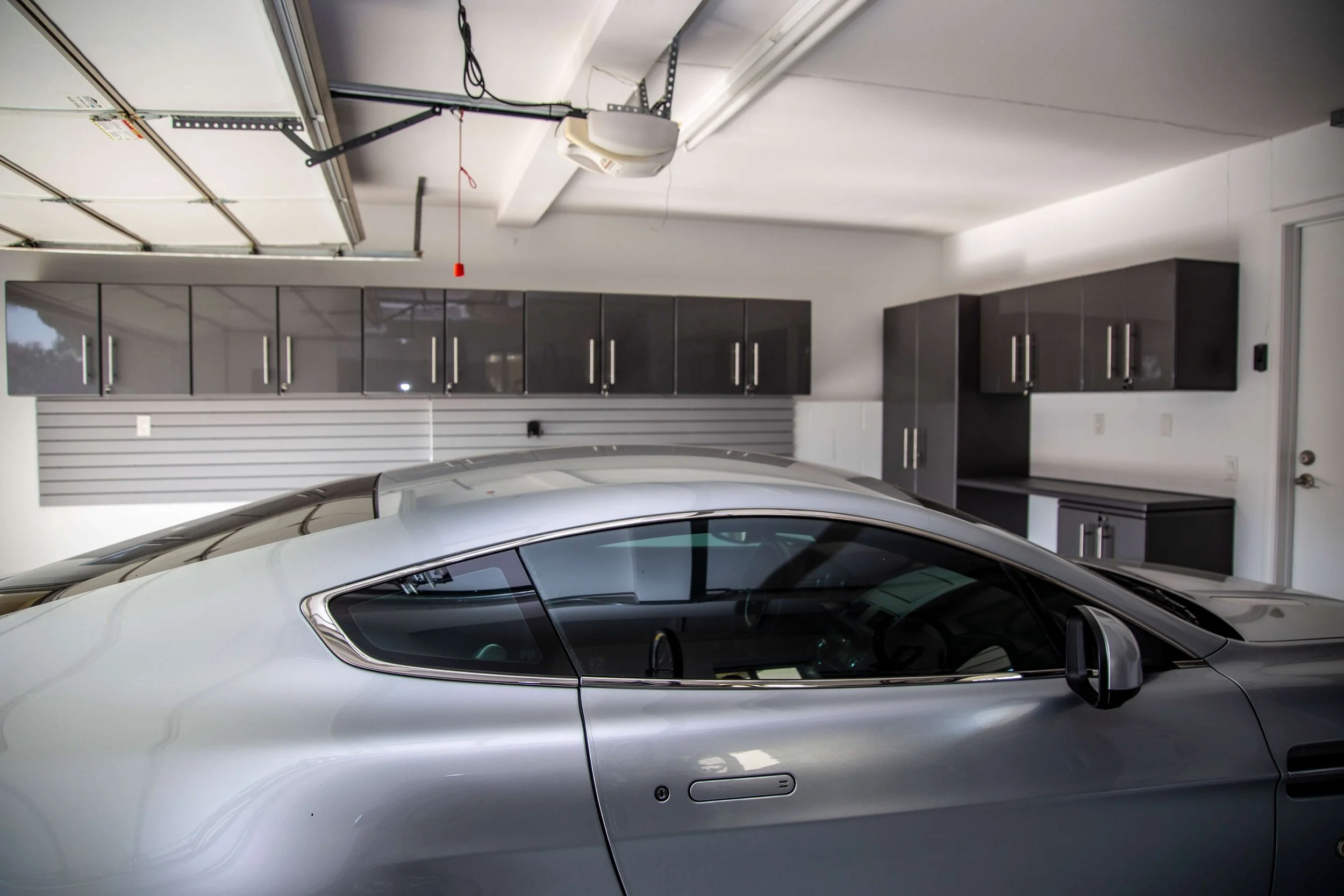 Interior of a modern garage with a silver sports car parked inside. The garage has dark gray cabinets and a white wall with horizontal slat paneling. An open garage door mechanism is visible on the ceiling.