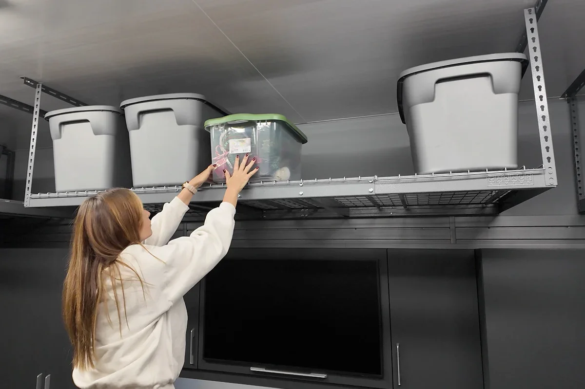 A woman reaching to place a plastic storage container on a metal shelf mounted on a gray ceiling in a room.
