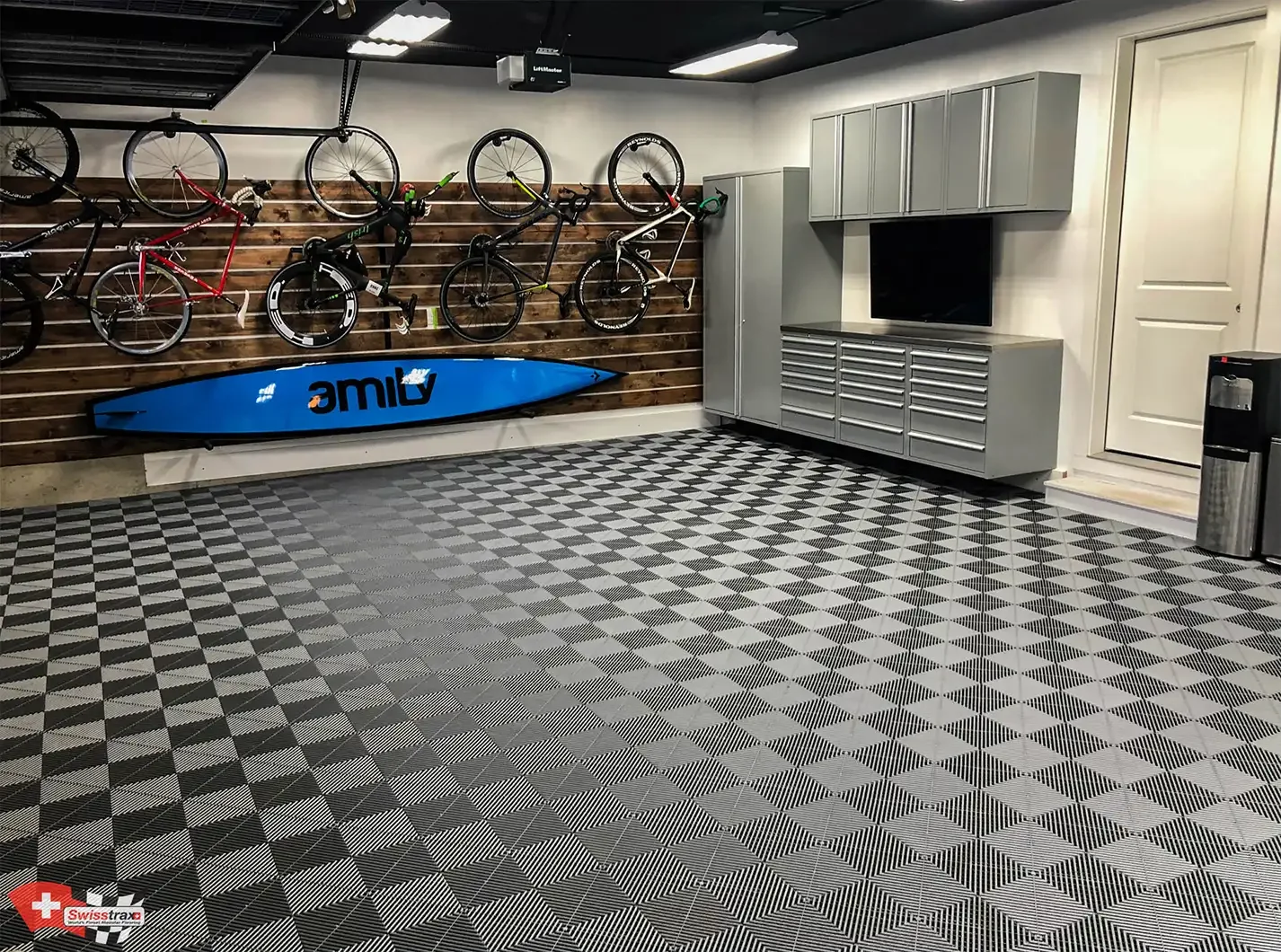 A garage or workshop with a wall of bicycles mounted on a wooden slat wall, a blue paddleboard beneath them, gray storage cabinets, a television, and a patterned black and gray floor.