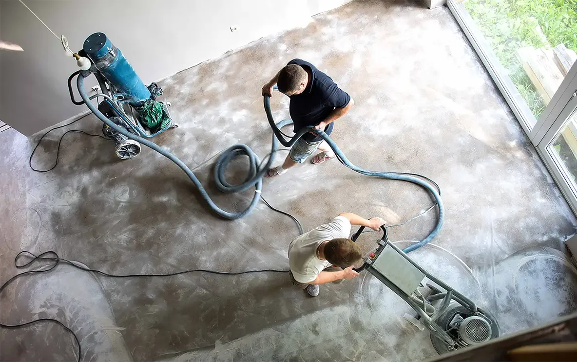 Two people are using floor sanding equipment to sand a concrete floor in a room with large windows.