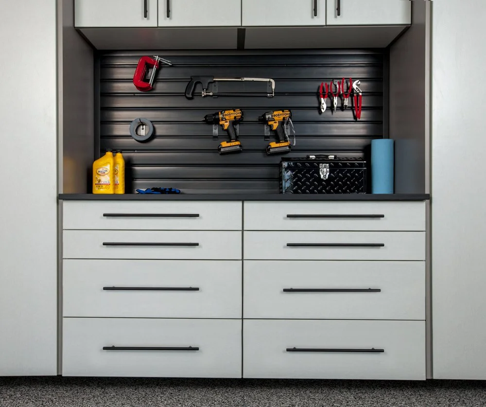 Garage wall with tools hanging, including a hacksaw, two cordless drills, a tape measure, pliers, and a cutter. On the countertop are two bottles of yellow cleaner, a pair of gloves, a black toolbox, and a rolled-up blue mat.