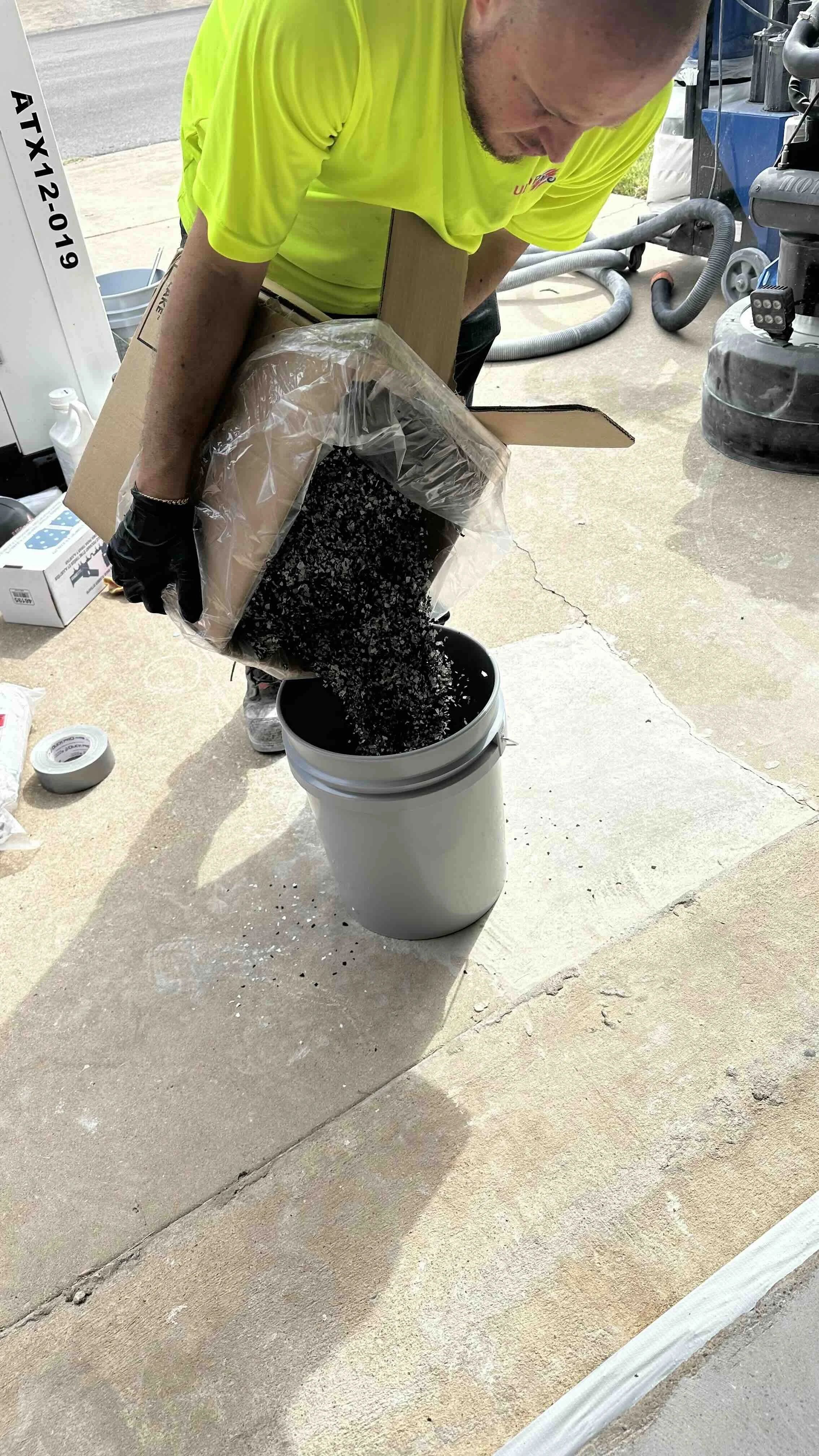 A person wearing a bright yellow shirt and black gloves pouring black and white speckled material from a large plastic bag into a white bucket outdoors. Various tools and equipment are visible in the background.