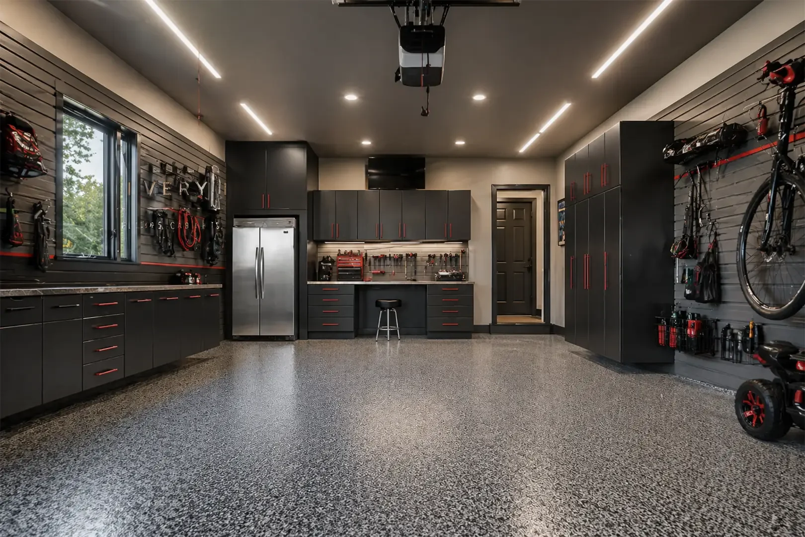 A clean, organized garage with black cabinets, a stainless steel refrigerator, pegboards with tools, bicycles on the wall, and a speckled epoxy floor.