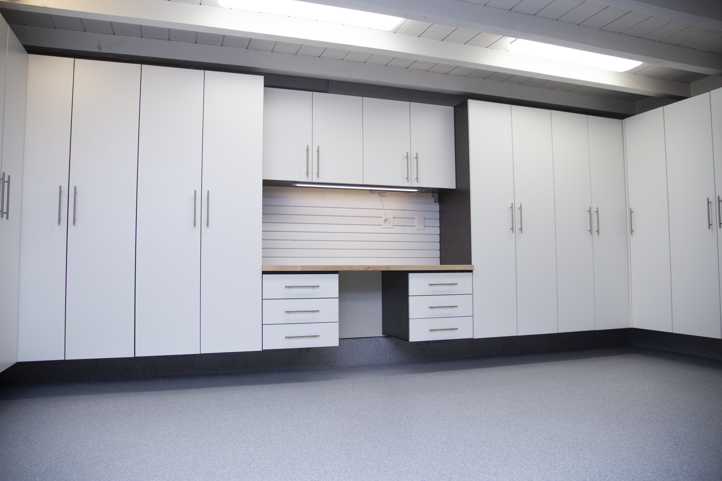 Interior view of a modern kitchen with white cabinets, a wooden countertop, and a gray wall with electrical outlets. The ceiling has a white wooden finish and a skylight, with gray carpeting on the floor.