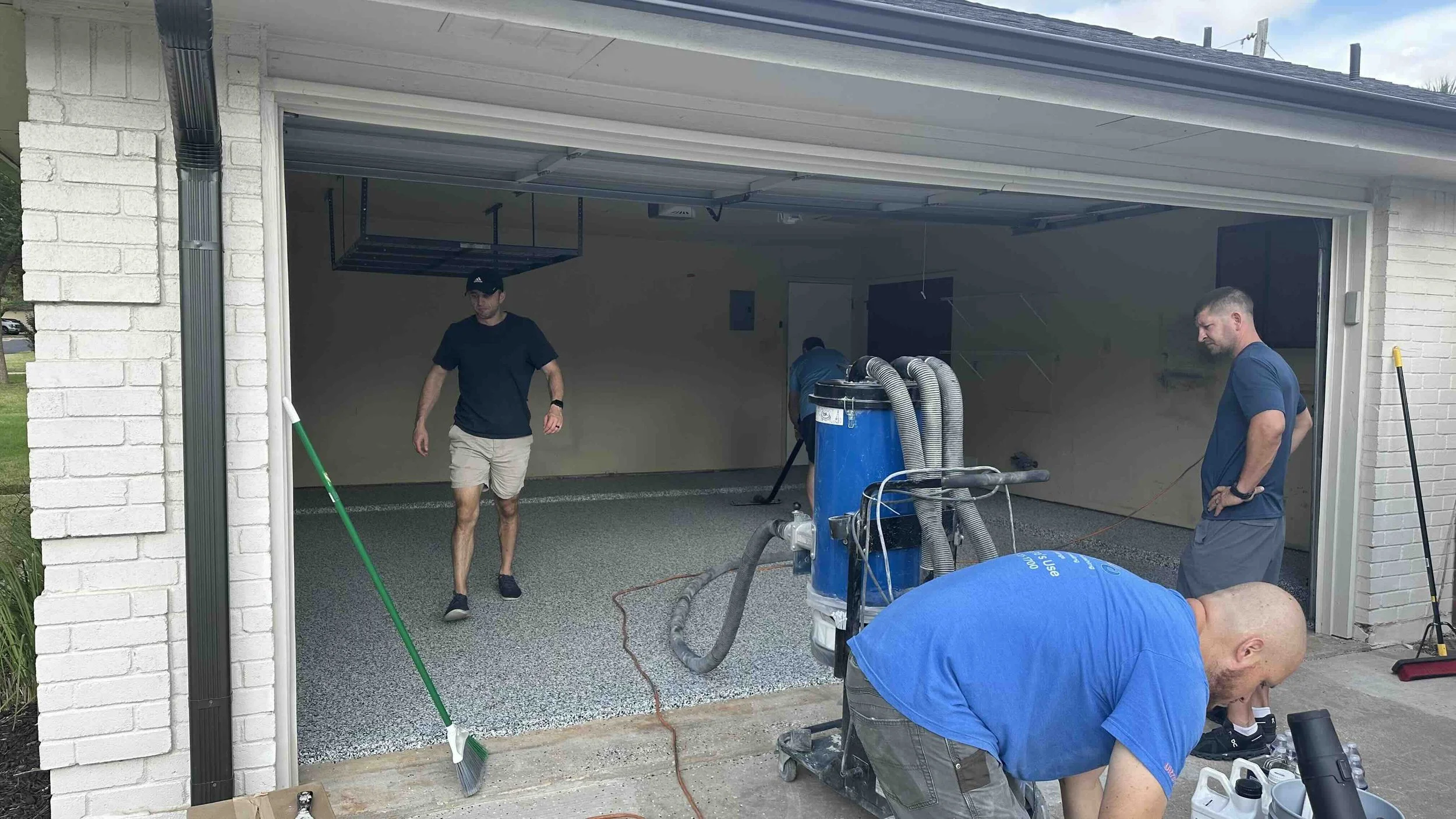 Men are installing or repairing a garage floor, using a vacuum or industrial machine, while others observe or prepare tools.