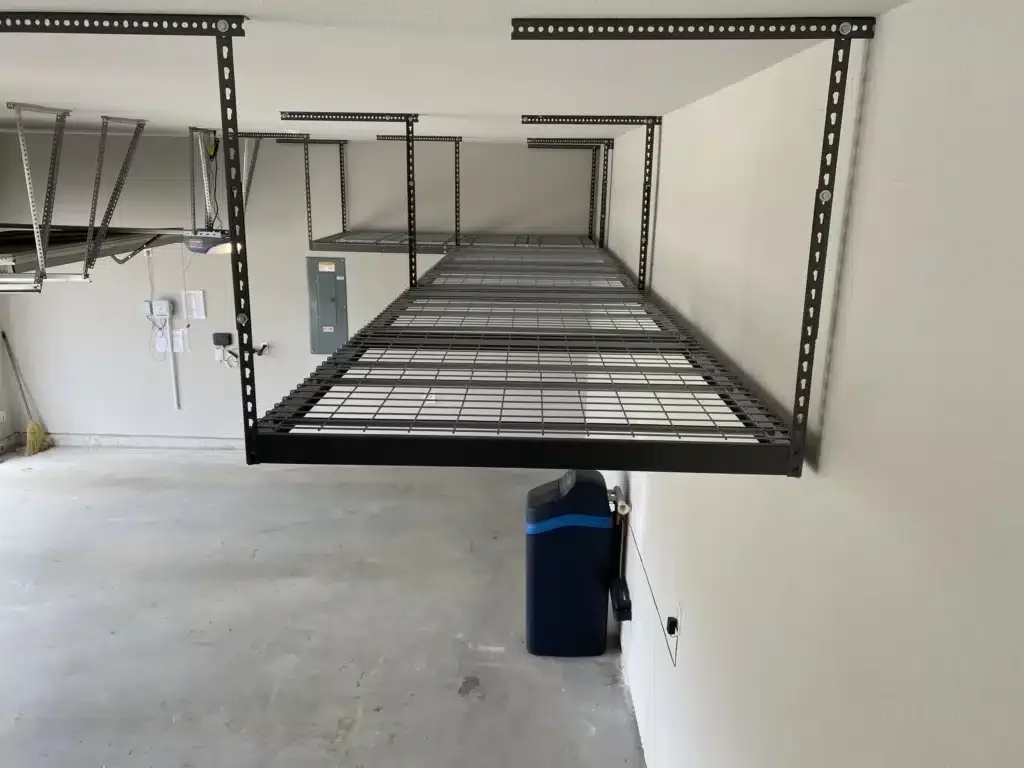 A black metal wall-mounted garage storage rack with no items on it, attached to a white wall in a garage, with a blue trash can below.